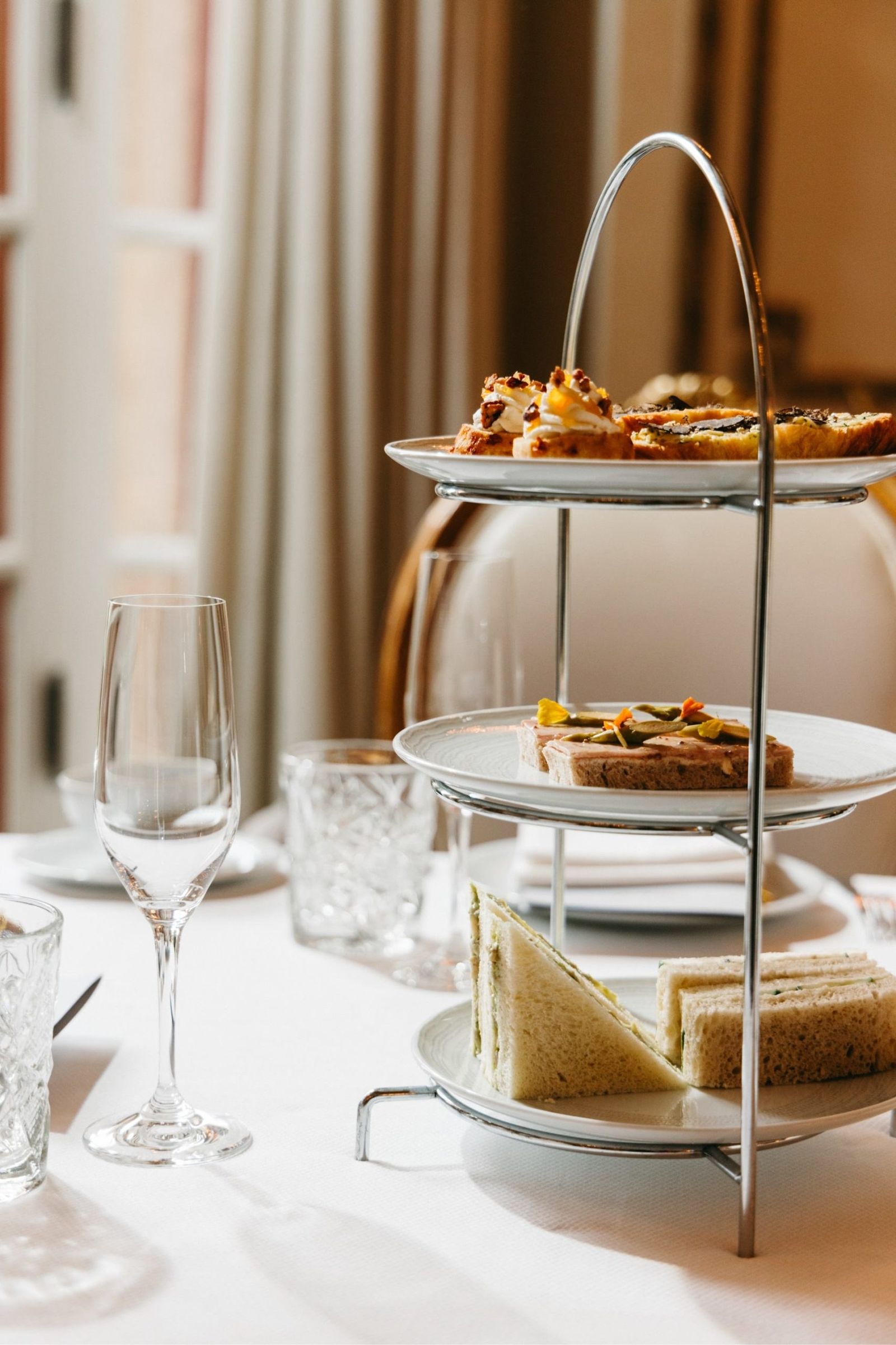 A three-tier stand of small pastries and sandwiches beside a champagne flute on a white tablecloth.