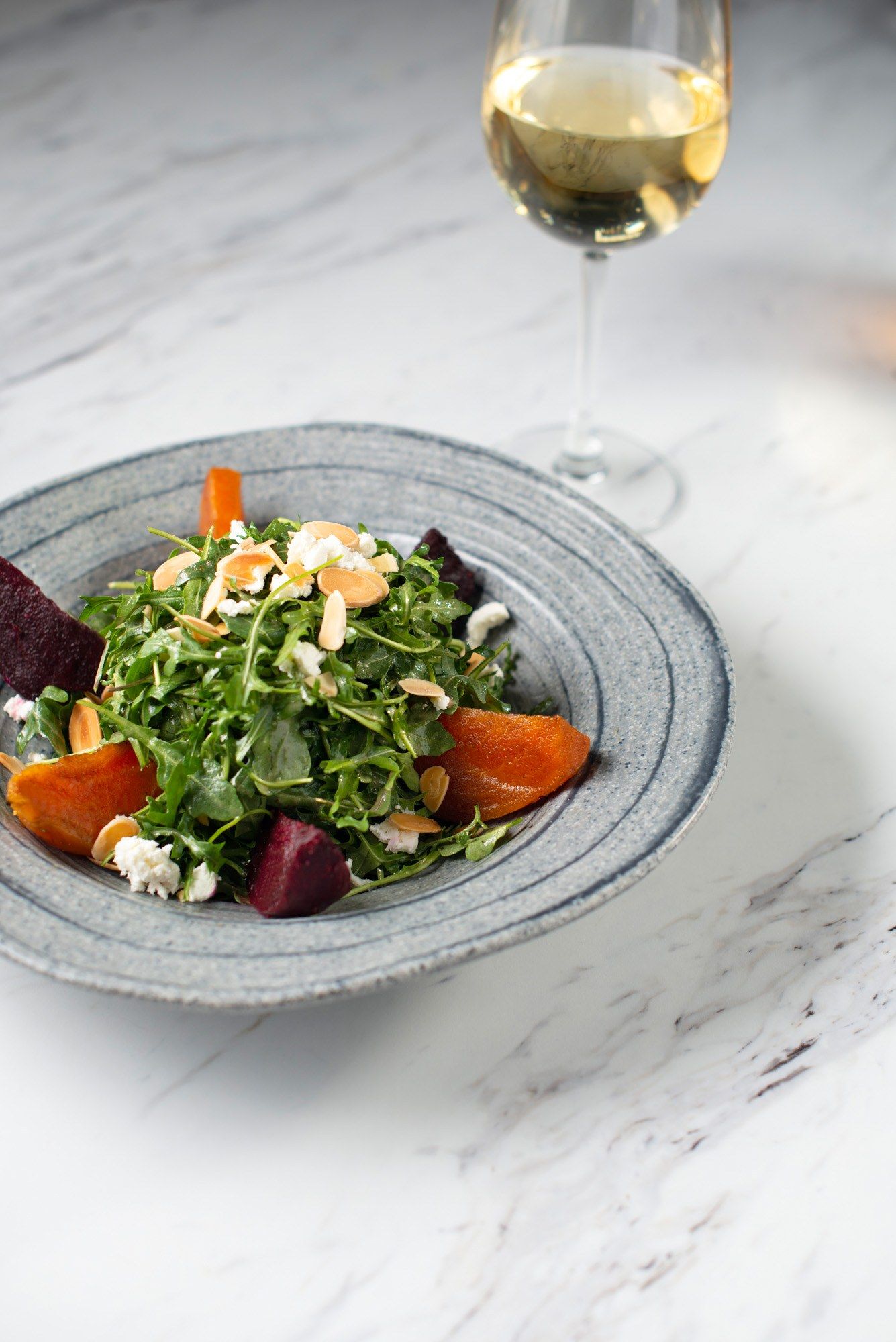 A salad of greens, beets and nuts in a gray bowl beside a glass of white wine.