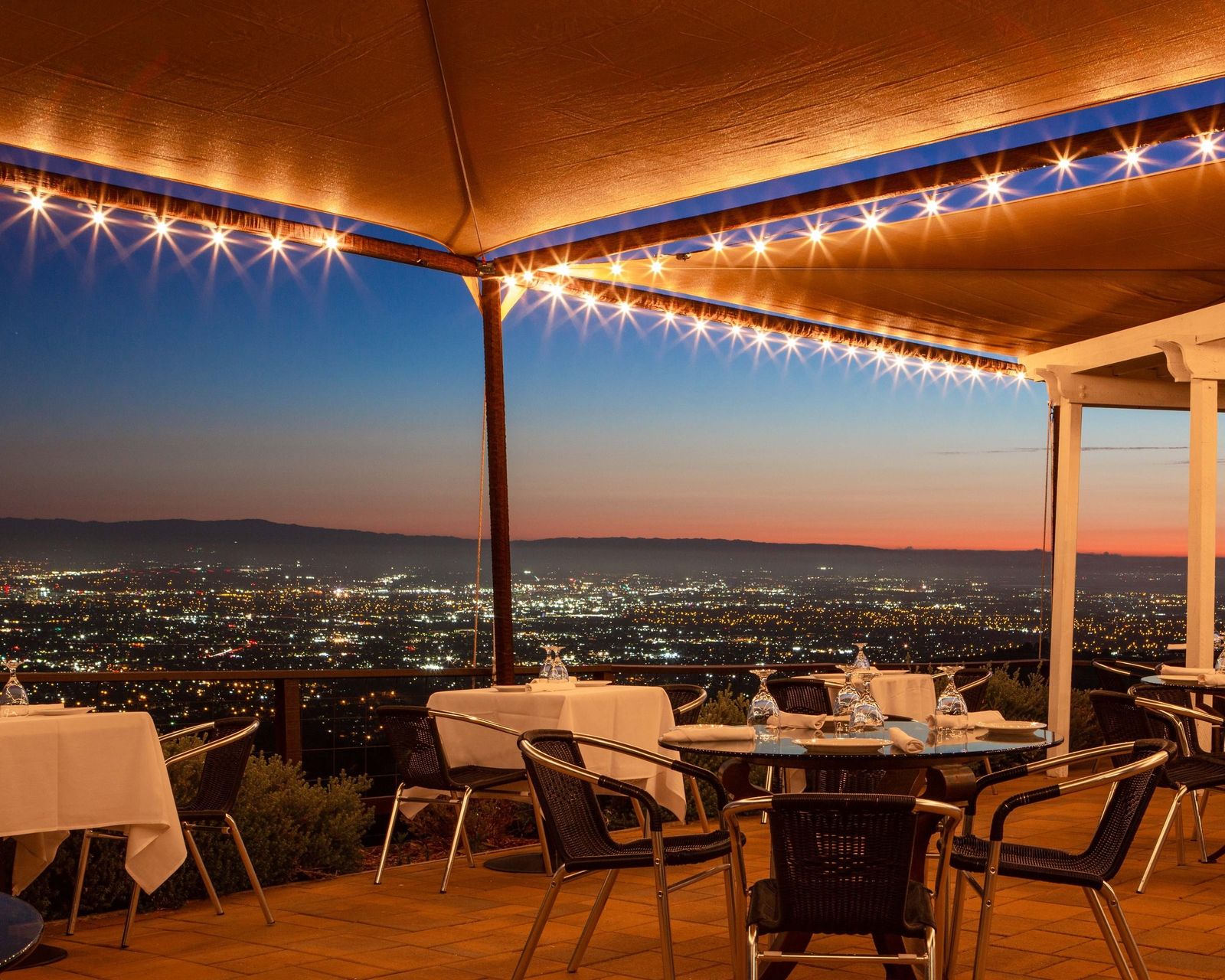 A covered hillside patio with string lights and set tables above the city lights at dusk