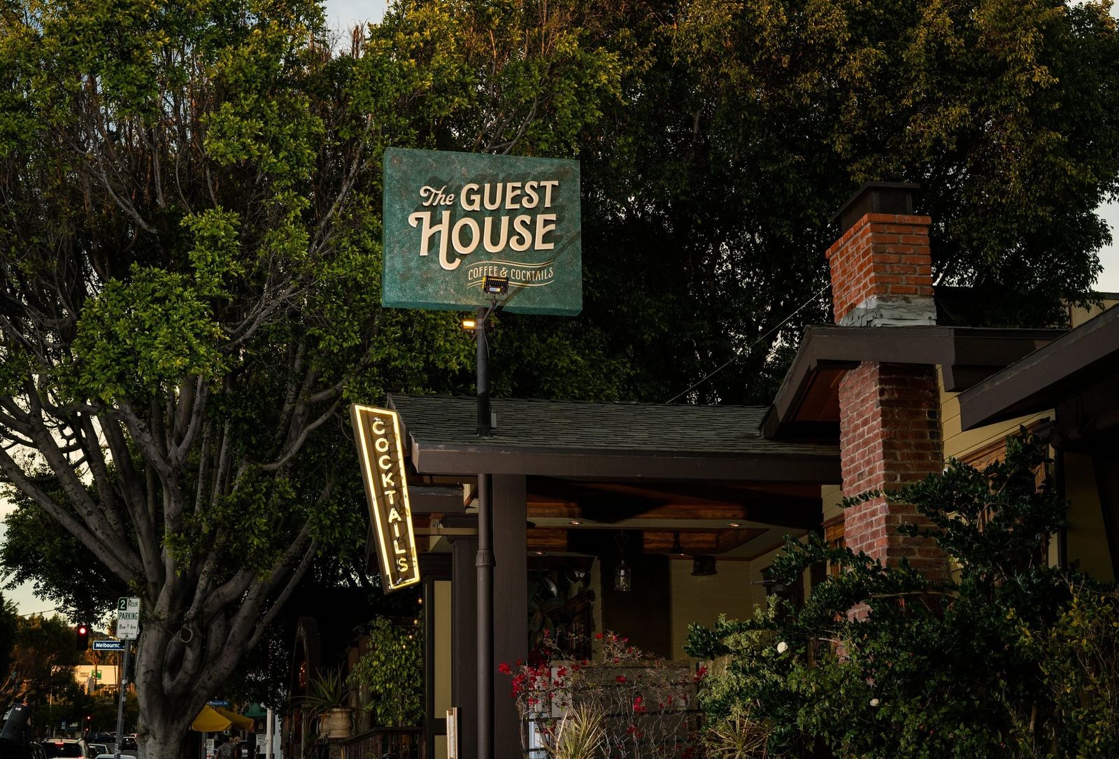 The Guest House's green sign among trees above a brick building, with an illuminated vertical Cocktails sign.