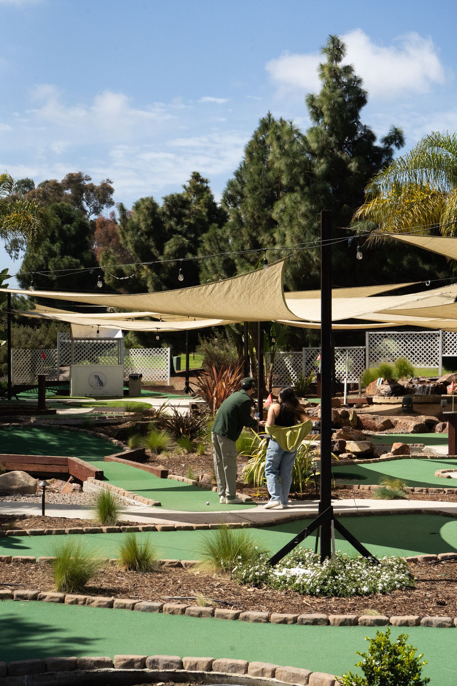 A mini-golf course with turf greens and sand traps under shade sails and tall trees.