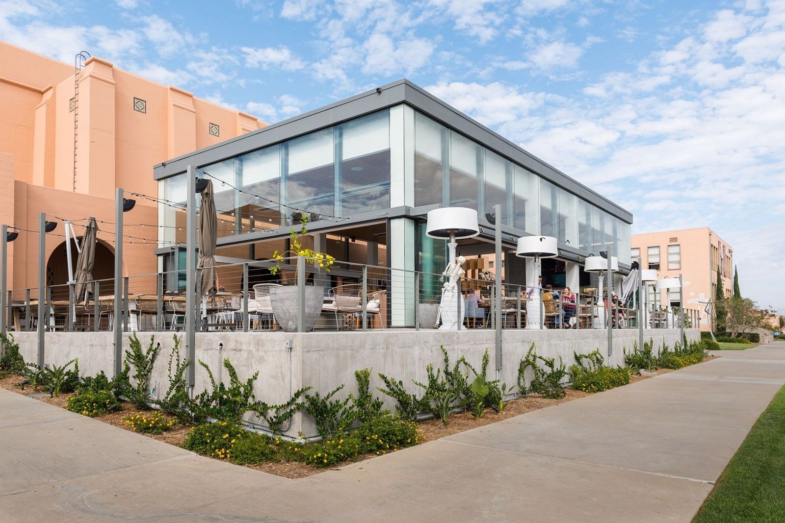 A glass-walled building with a pitched roof and outdoor patio on a corner, beside a path and landscaping under a blue sky.