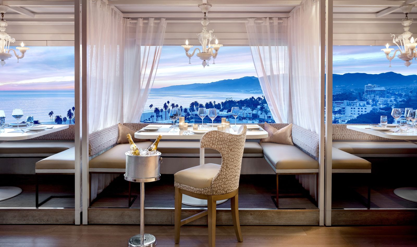 A dining room with banquettes and chairs along floor-to-ceiling windows overlooking the ocean and coast, with white drapes.
