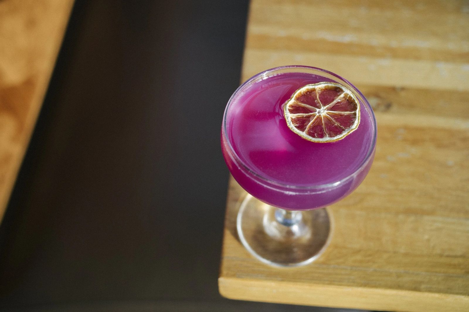 A purple cocktail with a dried citrus wheel in a coupe glass on a wooden board.