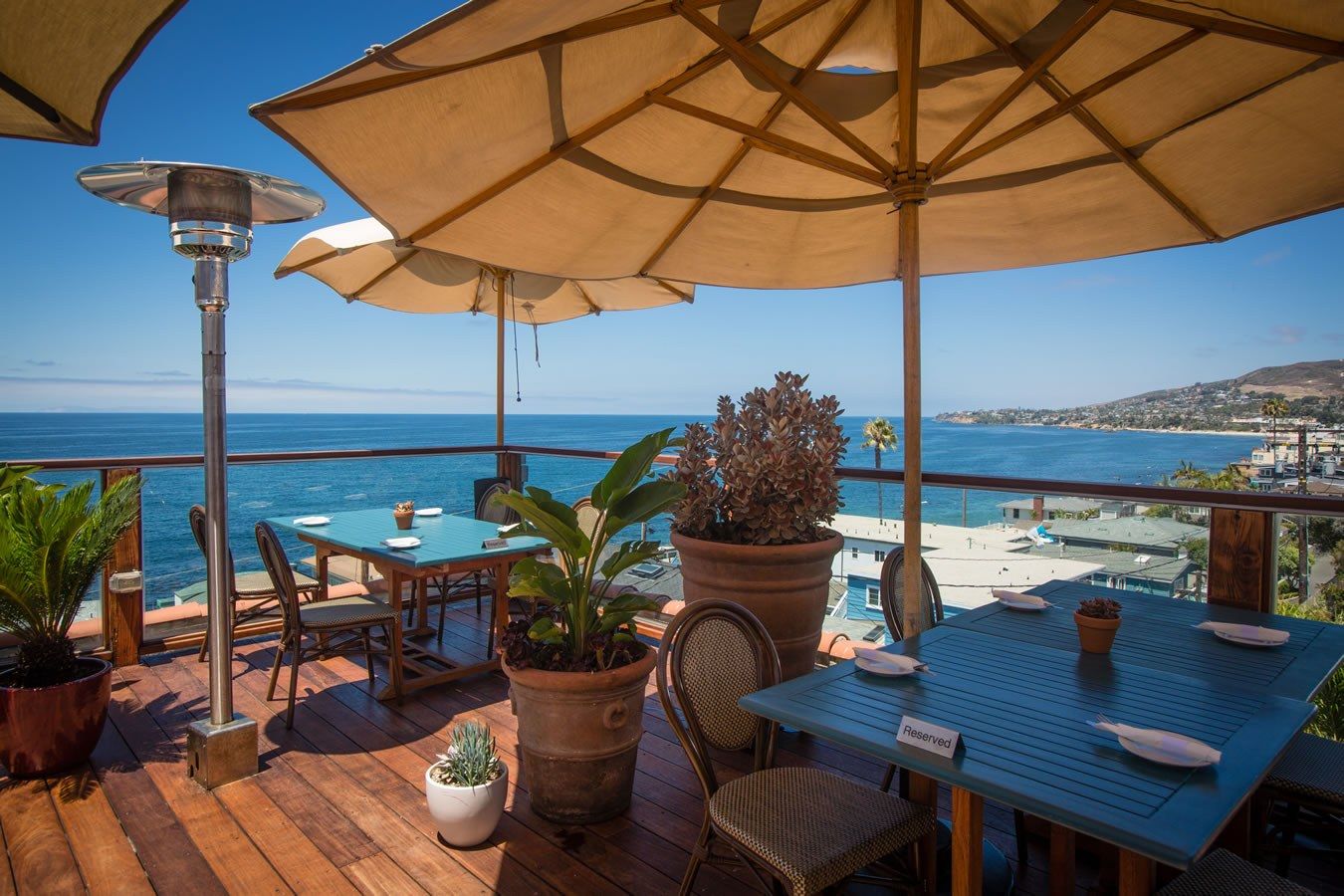 Blue tables and umbrellas on a wooden rooftop deck above the ocean.