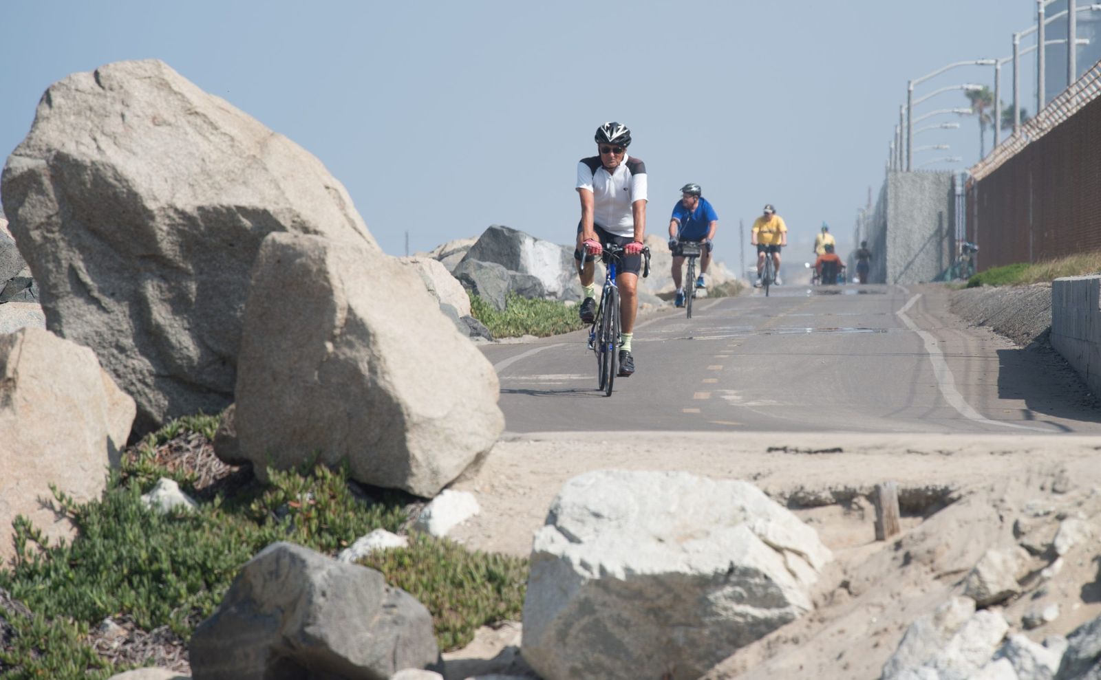 Cyclists on a paved beach path beside large boulders under a hazy sky.