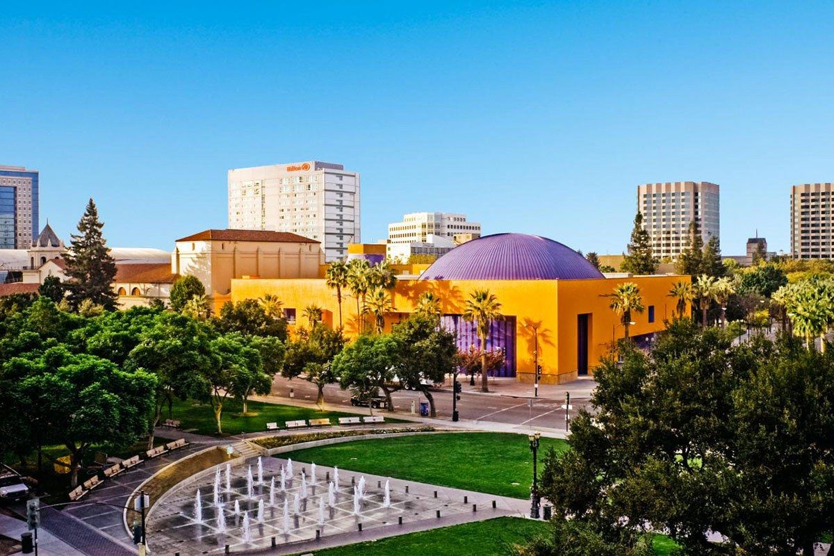 Exterior of the Tech Interactive's orange building with a purple dome, fountains and a lawn in the foreground