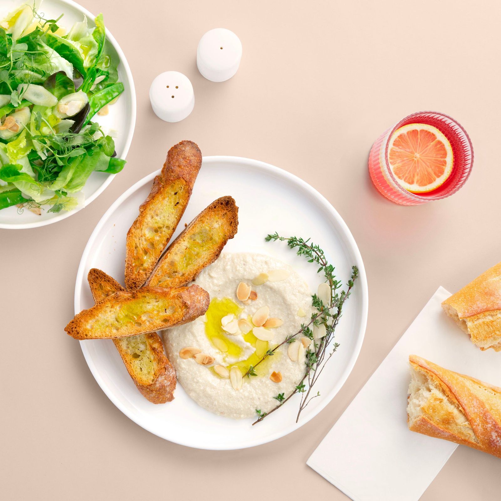 A plate of hummus with grilled bread and herbs beside a grapefruit drink and a salad.