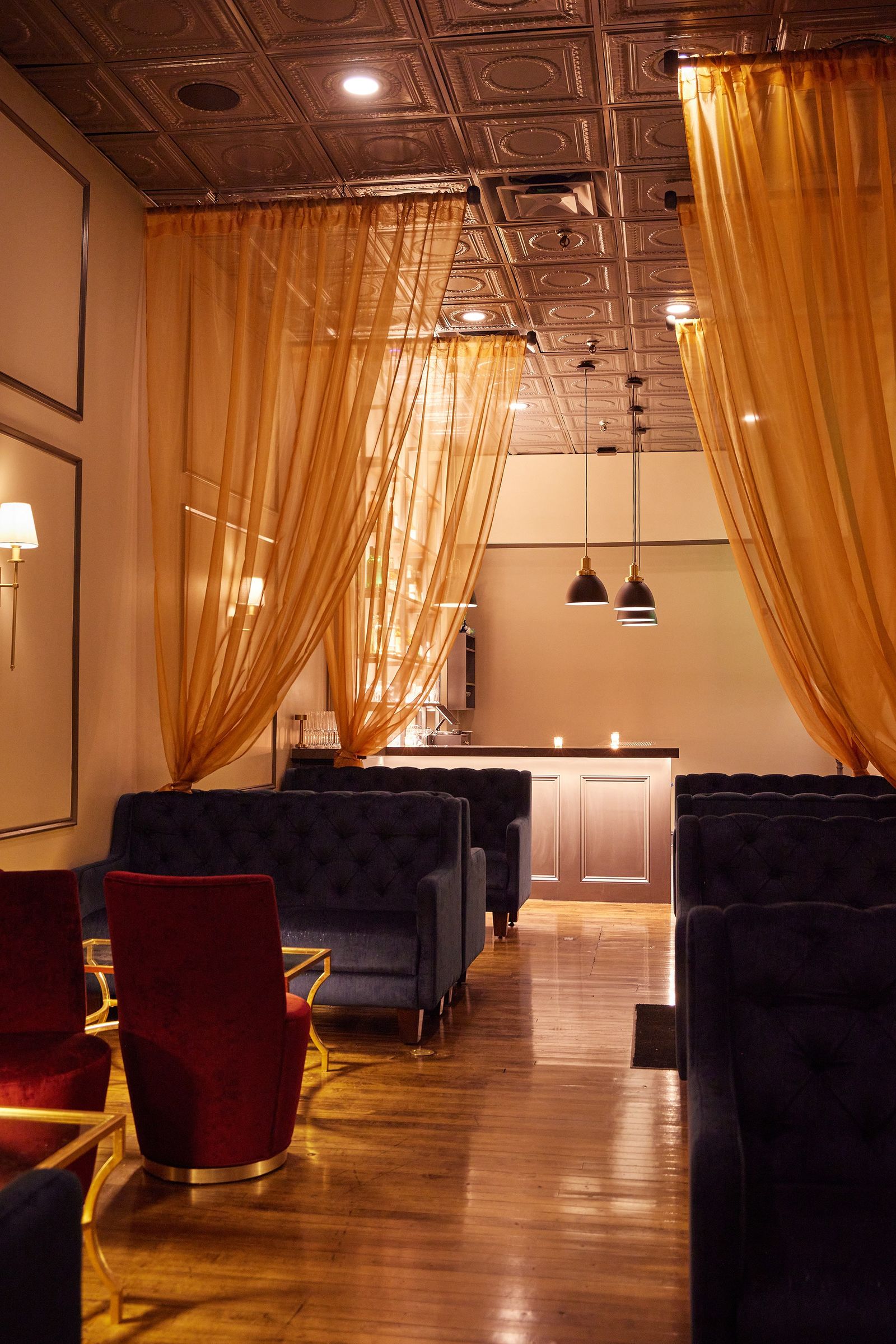 A dim lounge with golden curtains, navy booths and red armchairs