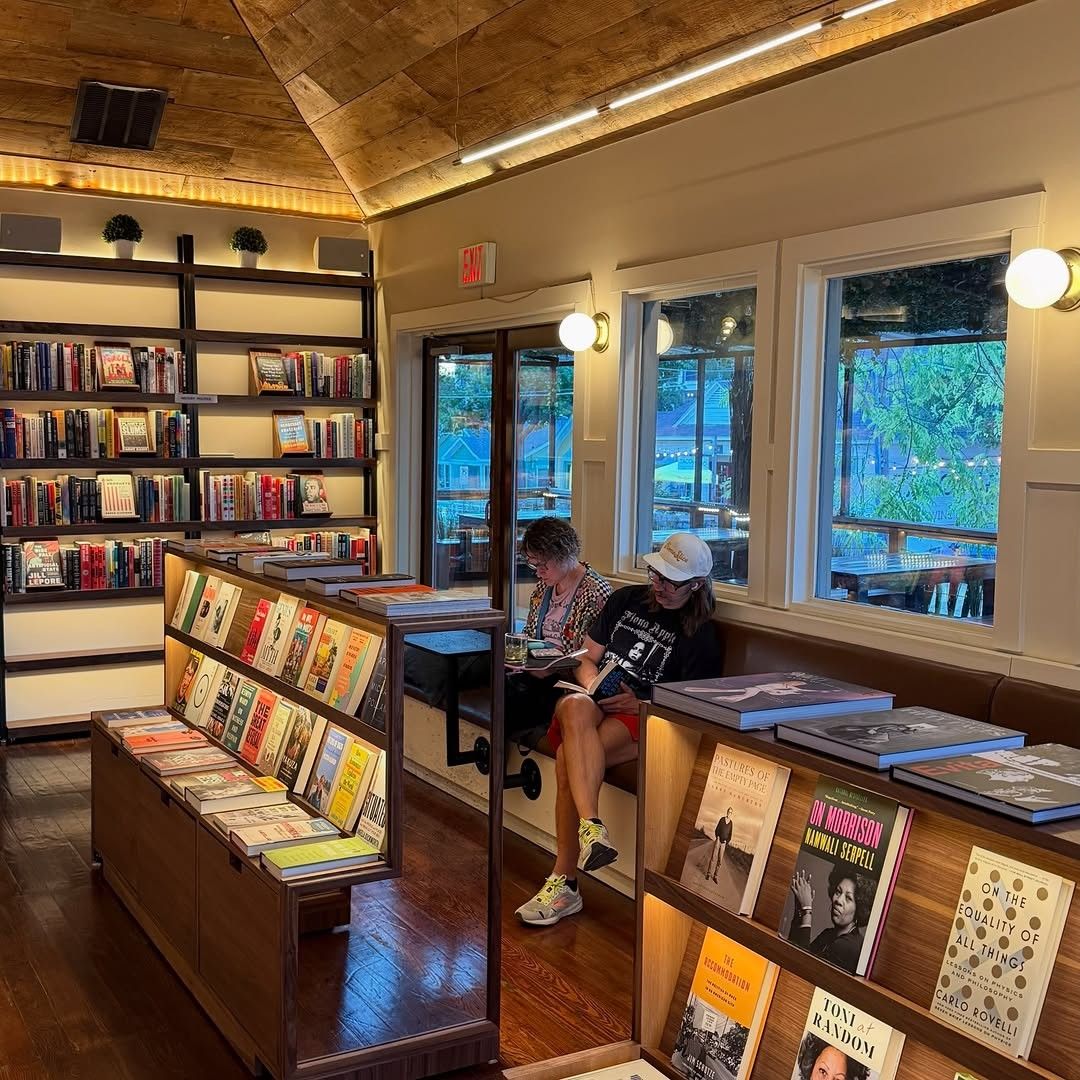 A bookshop with wooden shelves of books, display tables of titles and windows onto trees.