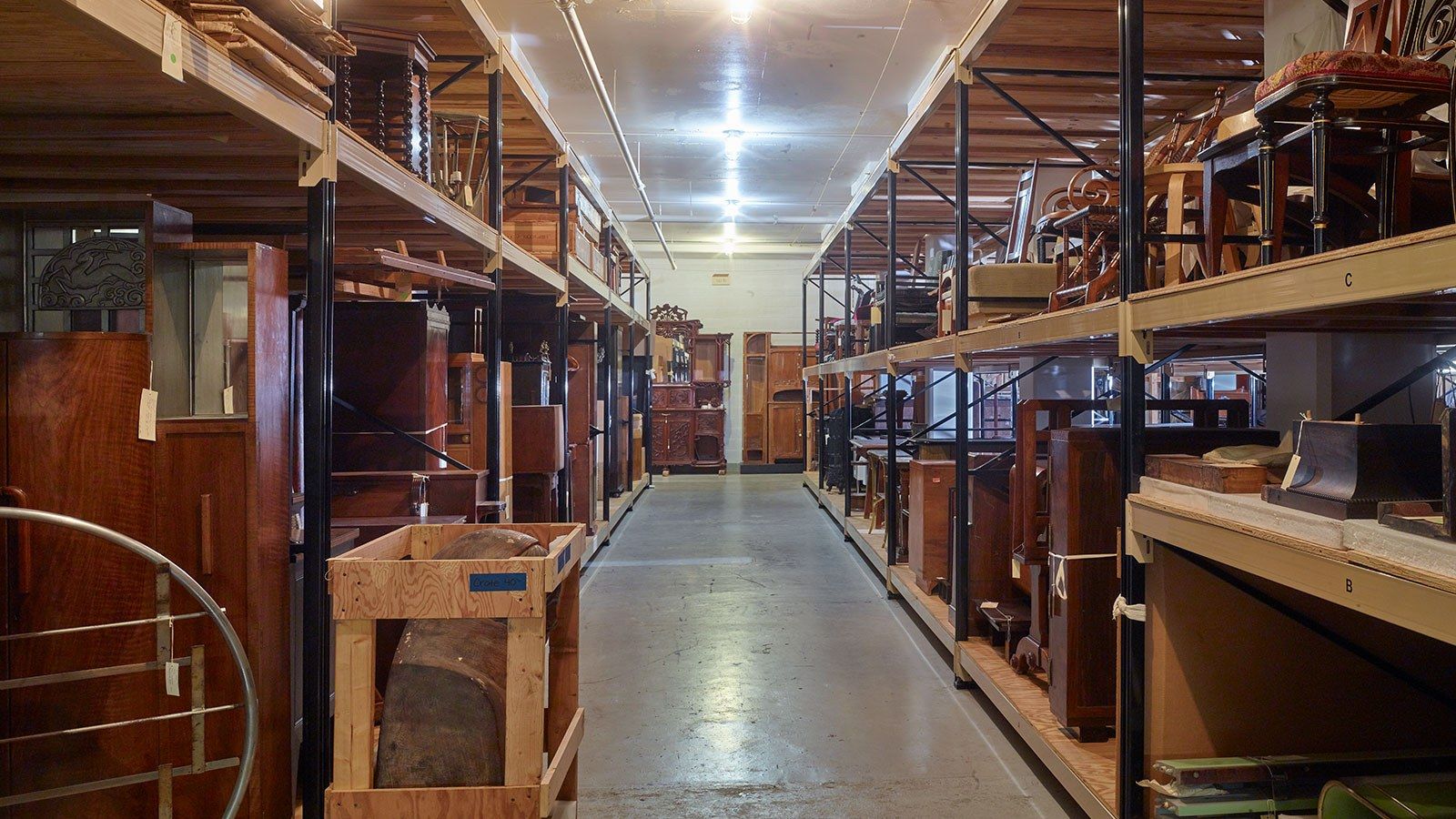 Long aisle between tall shelving holding wooden furniture and collection objects