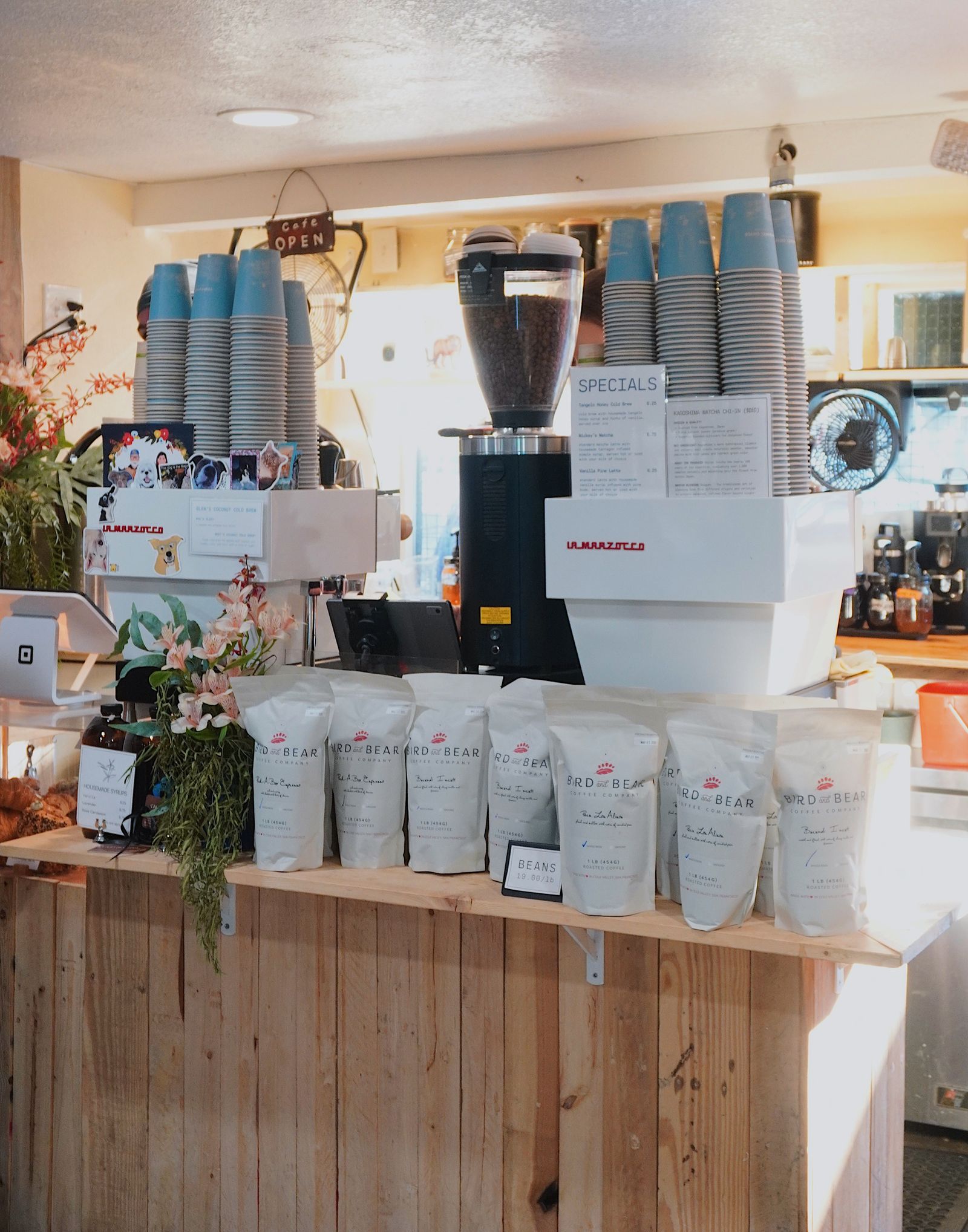 A wooden coffee counter with an espresso machine, stacked blue cups, bags of beans and a vase of flowers.