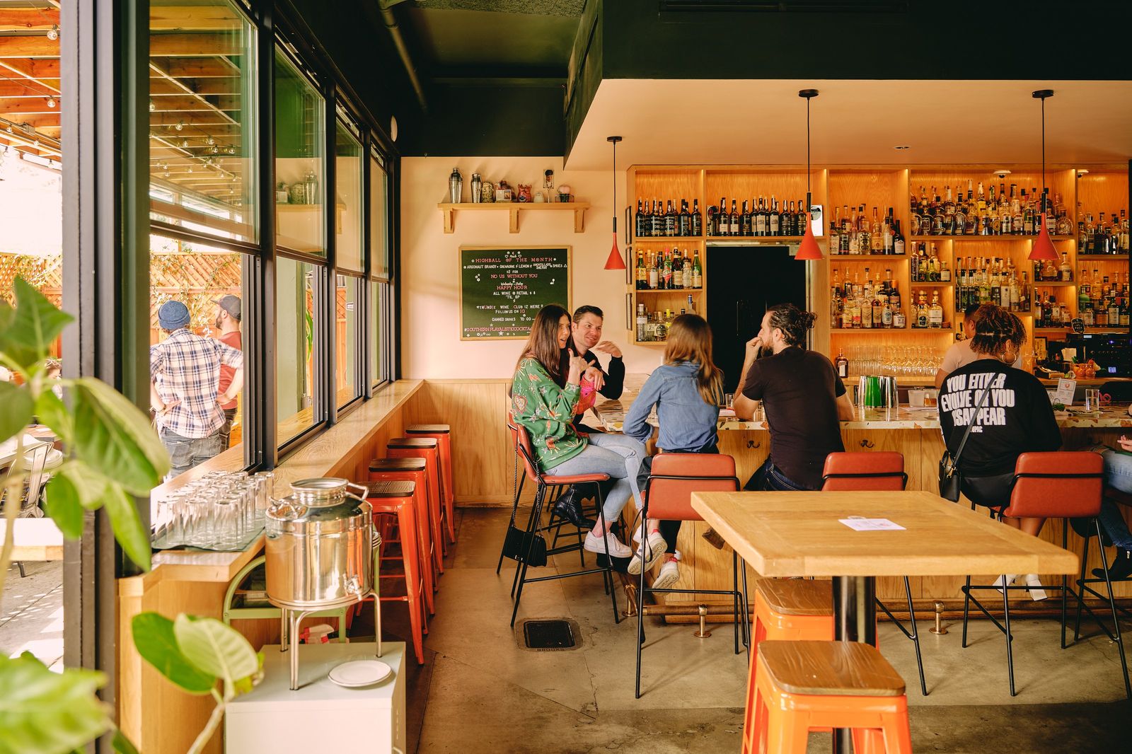 Bright dining room with a long bar, orange stools and shelves of bottles seen through tall windows