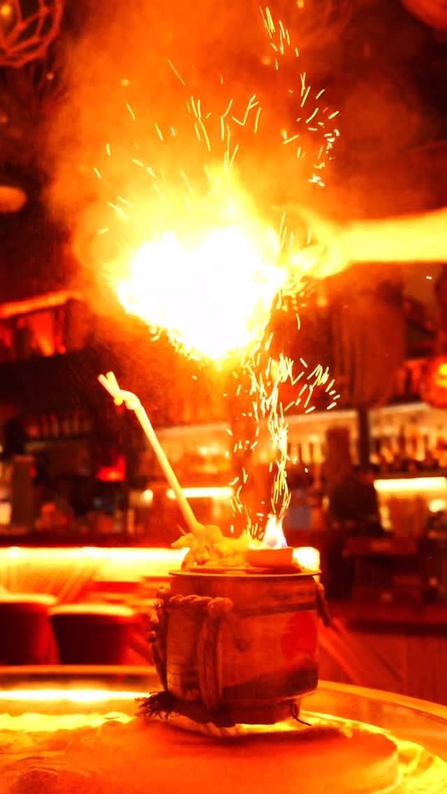 A flaming tiki drink throwing sparks above a ceramic mug in a dim, orange-lit bar.
