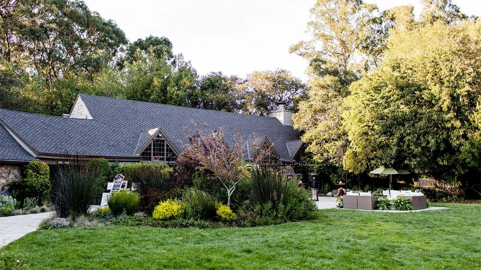 A gray-roofed lodge with a lawn and flowering garden beds under trees.