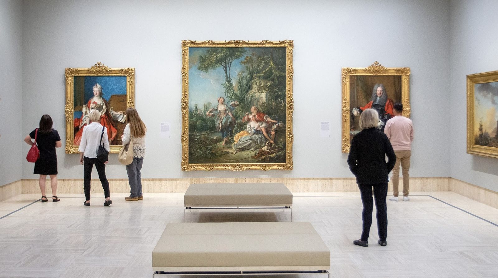 A bright gallery with framed paintings on pale walls and a few visitors looking at them.
