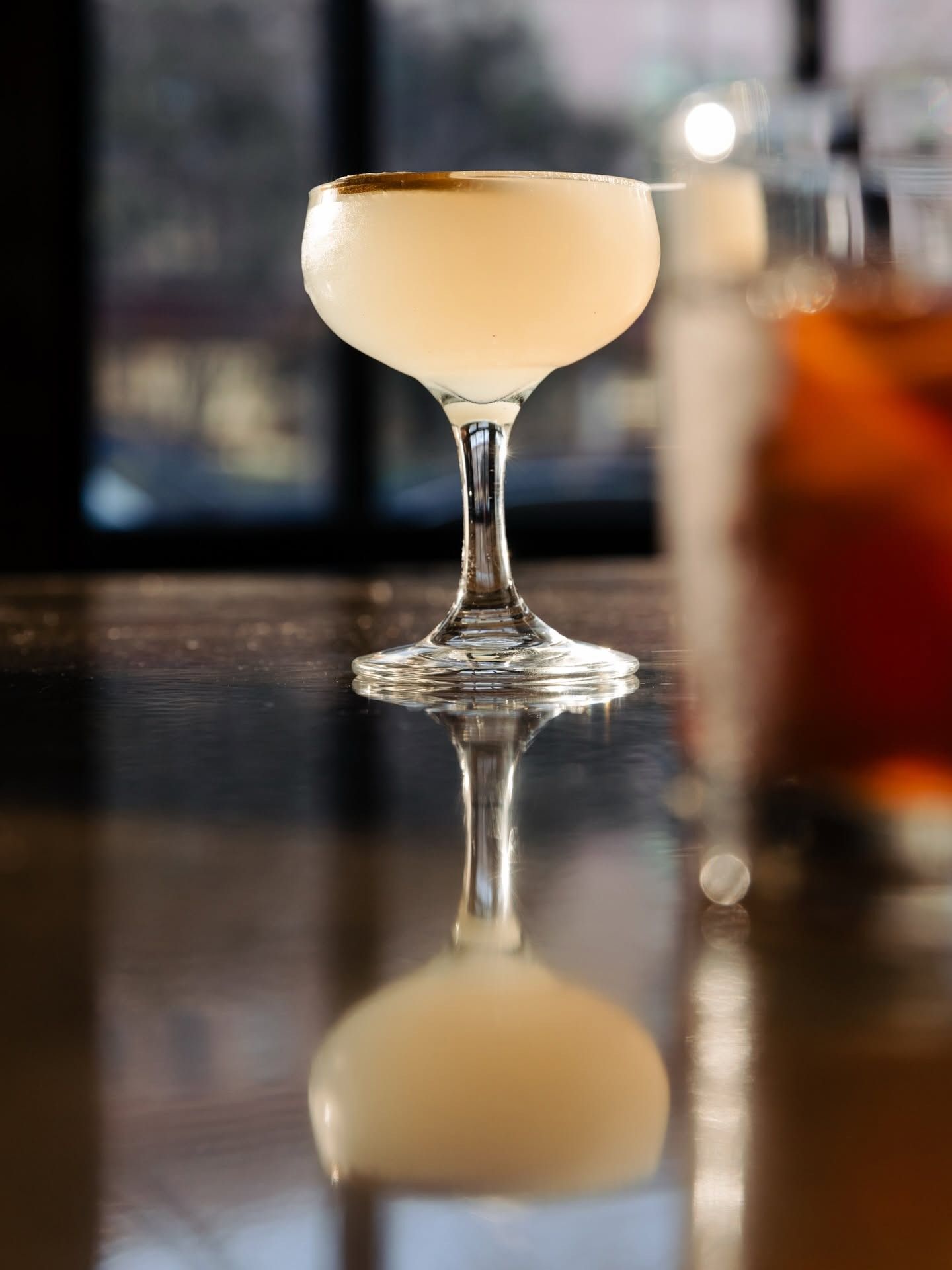 A coupe glass with a pale pink cocktail, reflected in a dark bar top.