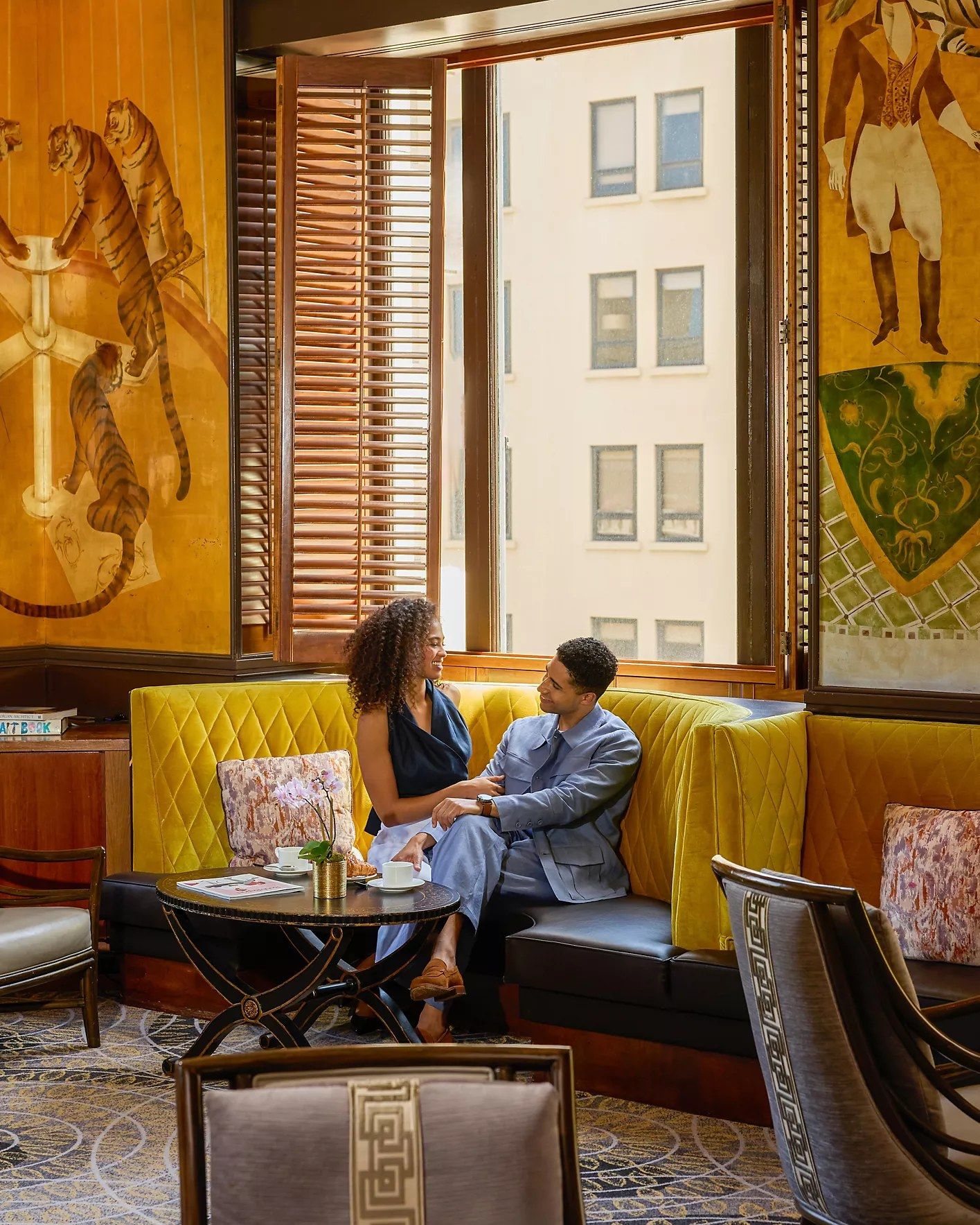 Two people seated on a yellow booth by a window in a room with gold tiger murals.