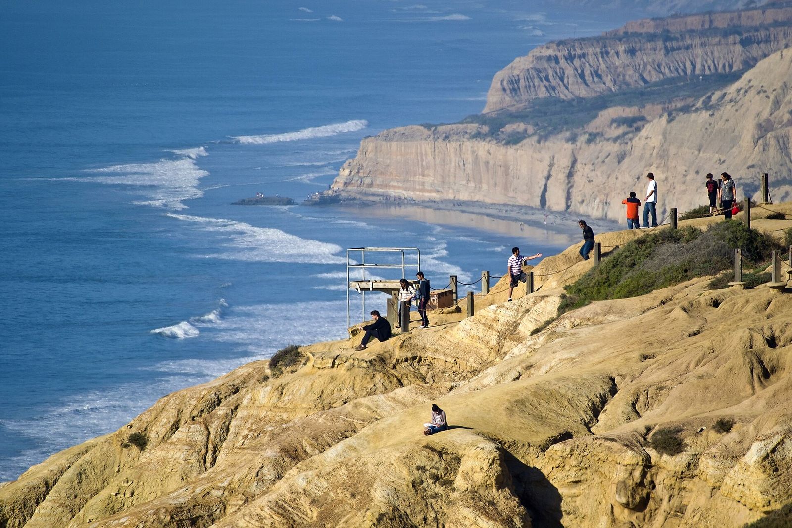 Sandstone cliffs above the ocean with a few visitors at a lookout and surf below.
