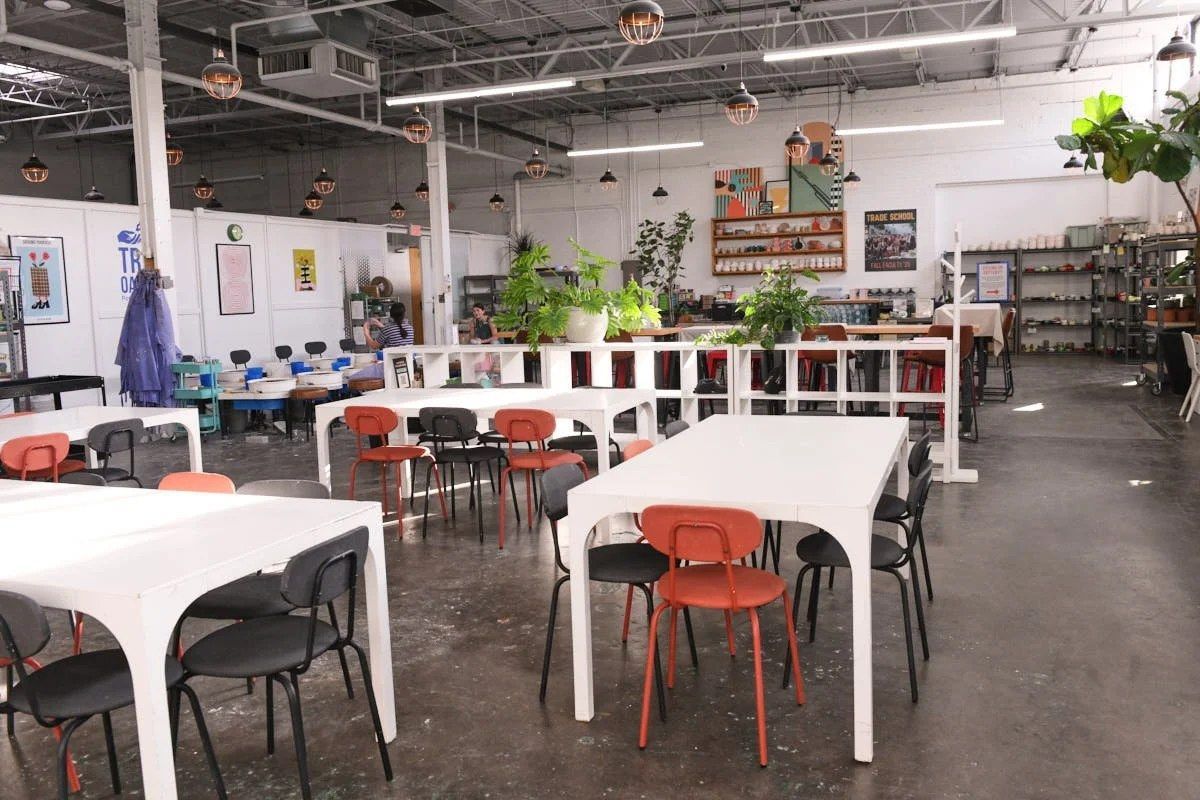 A bright studio workshop with long white tables and orange chairs, shelves of supplies and tools along the back wall.
