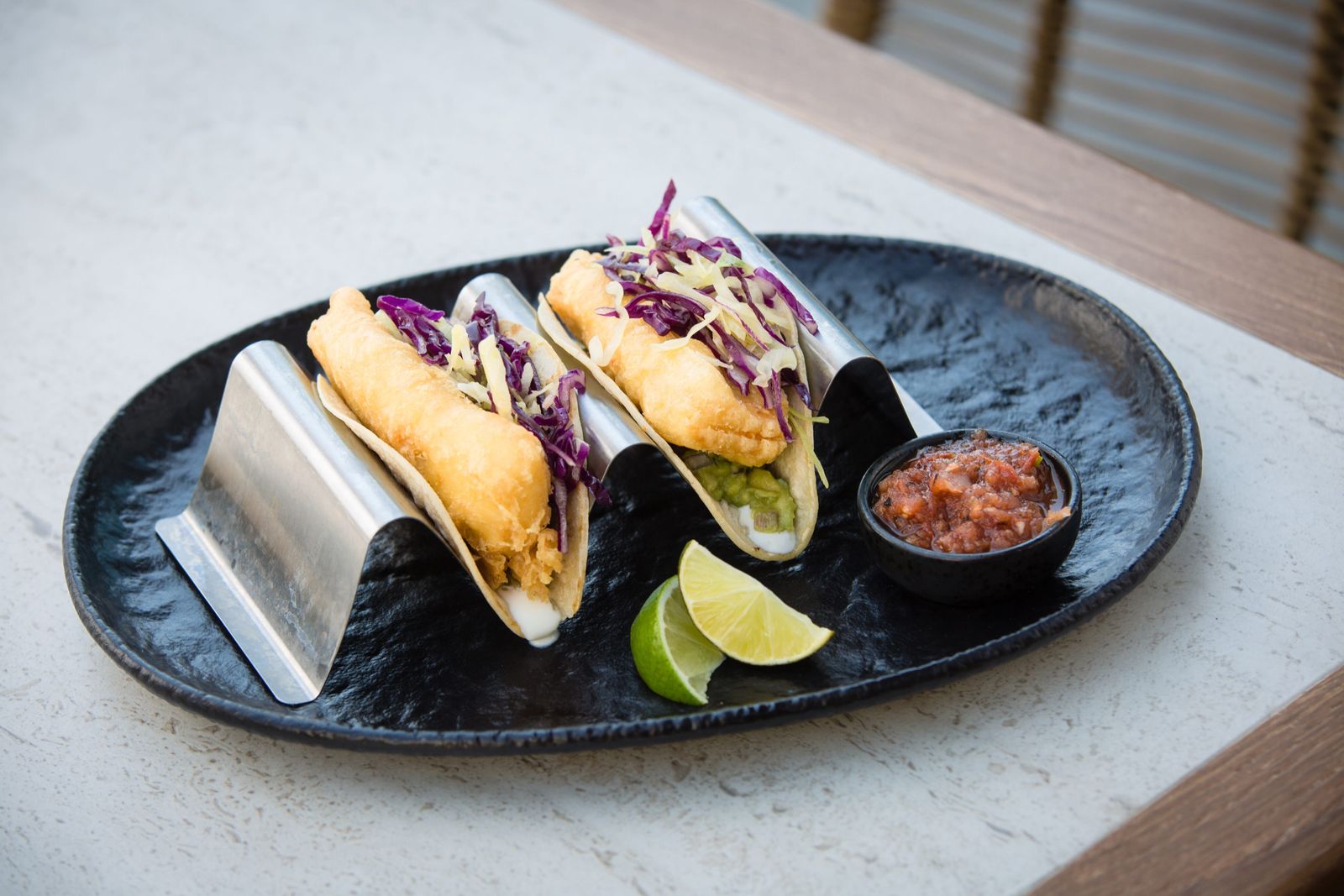 Two fried tacos in a metal holder on a dark oval plate with a lime and a dish of salsa.