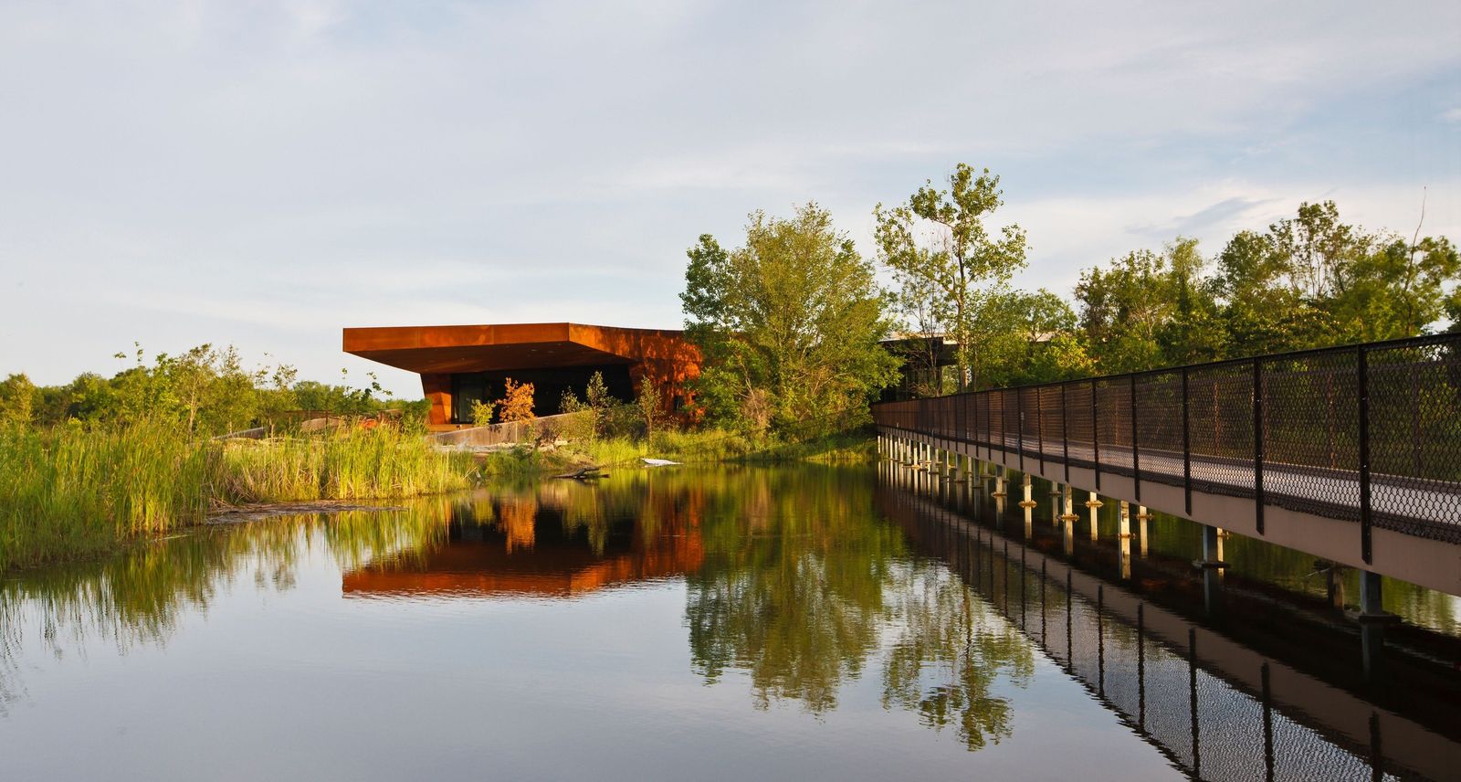 A rust-colored nature center building beside a long boardwalk, both reflected in a still pond with trees behind.