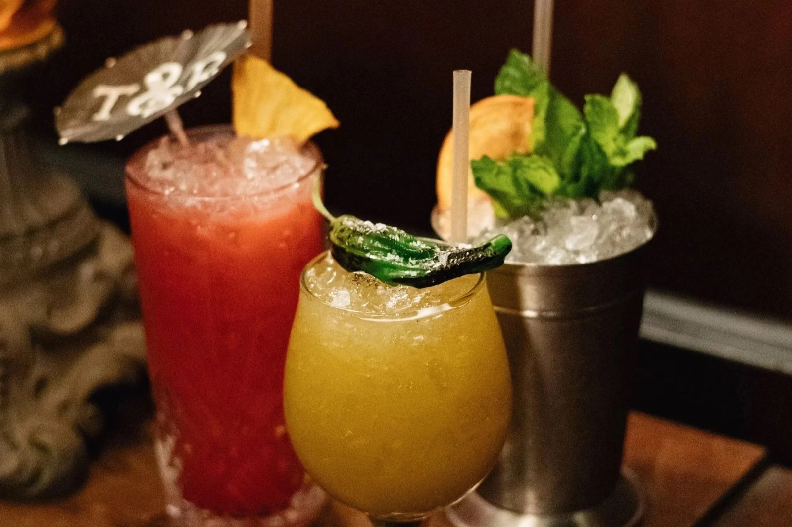 Three cocktails, one red, one yellow and one in a metal cup with mint, on a dark bar.