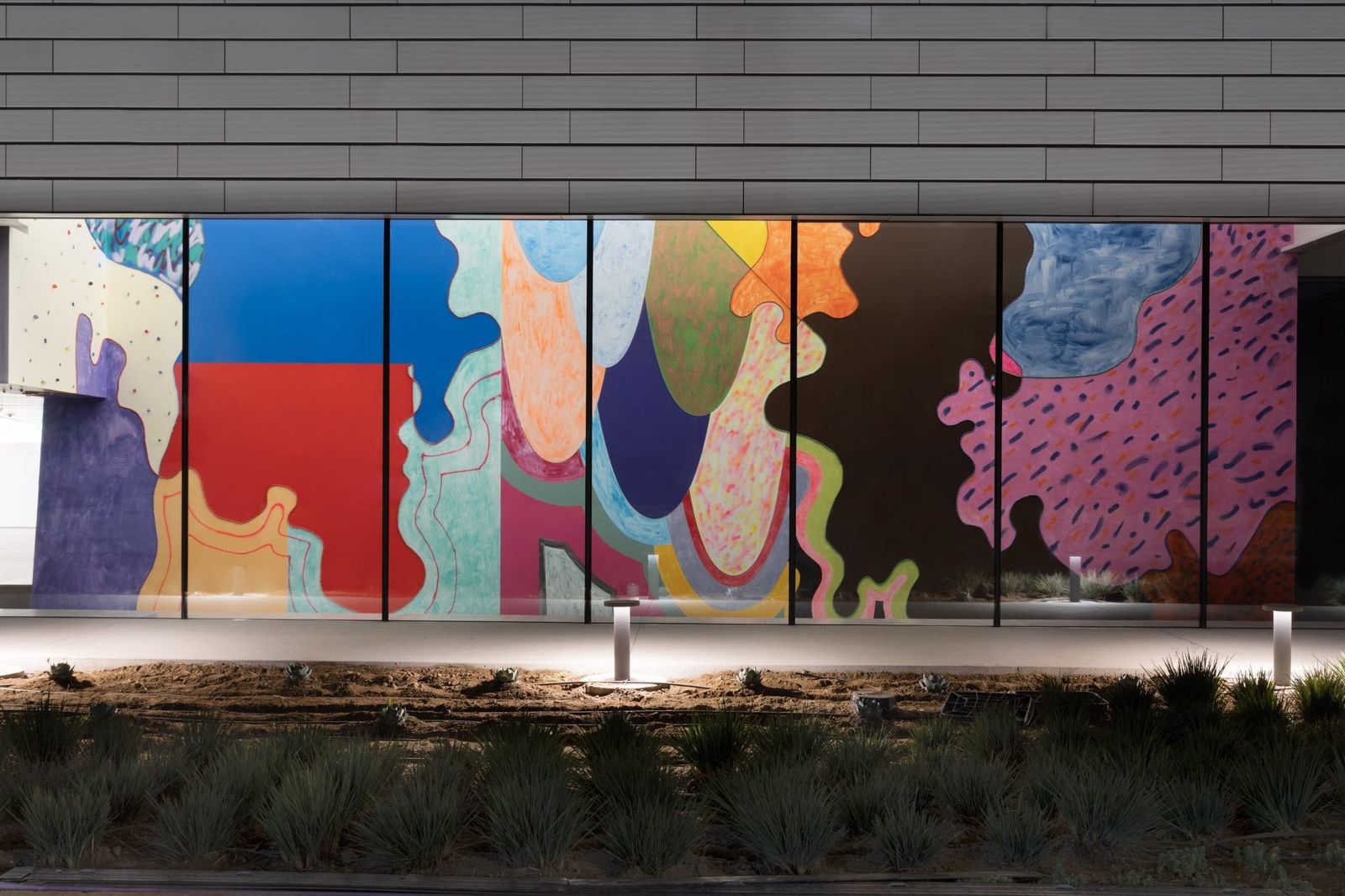The museum's dark, textured facade with a colorful abstract mural panel and low plantings in front.