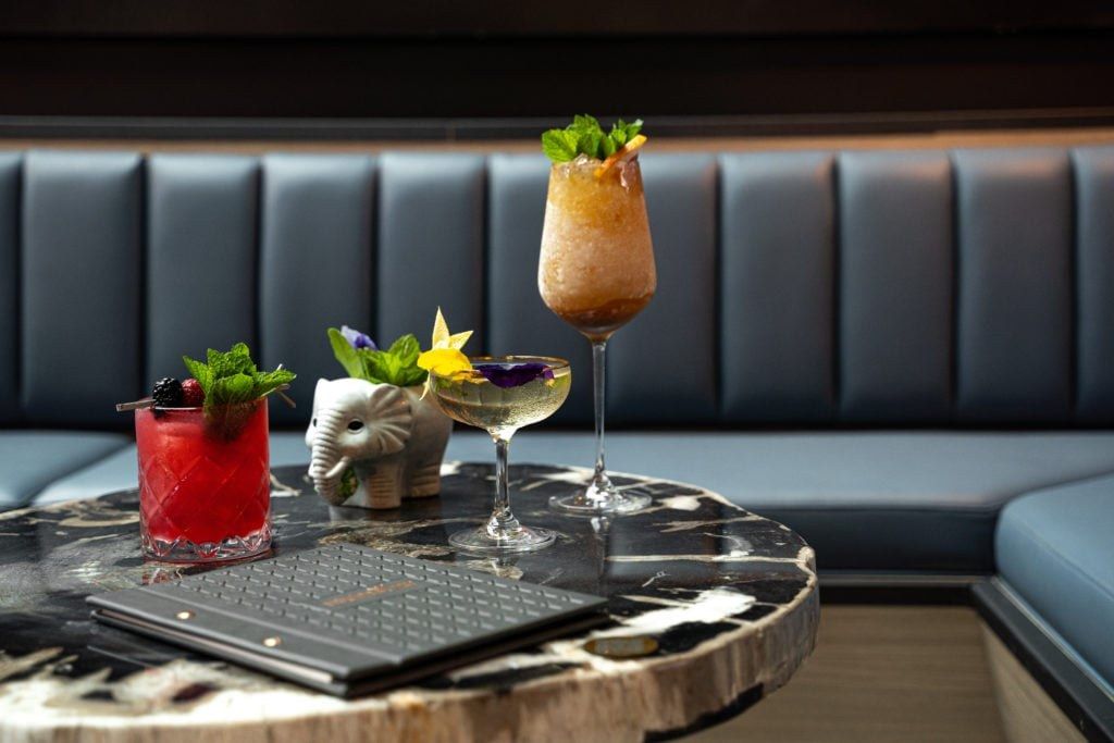 Three colorful cocktails on a round marble table in front of a blue tufted banquette.