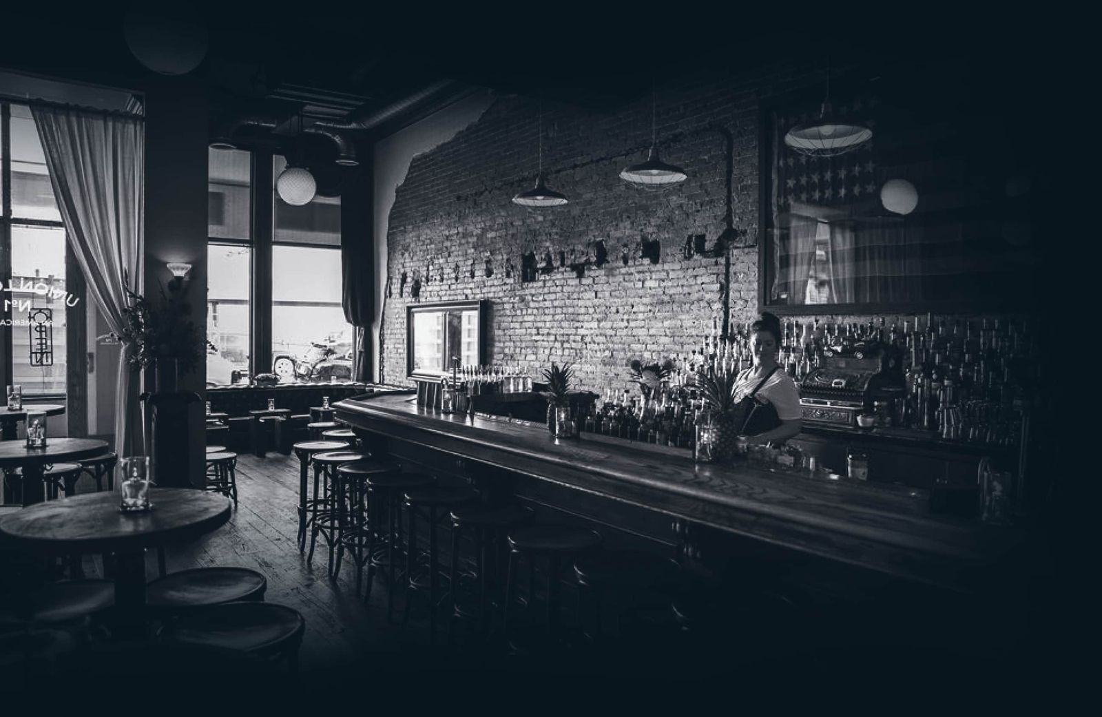 A dim long bar with stools and a brick wall in a room lit through tall windows.