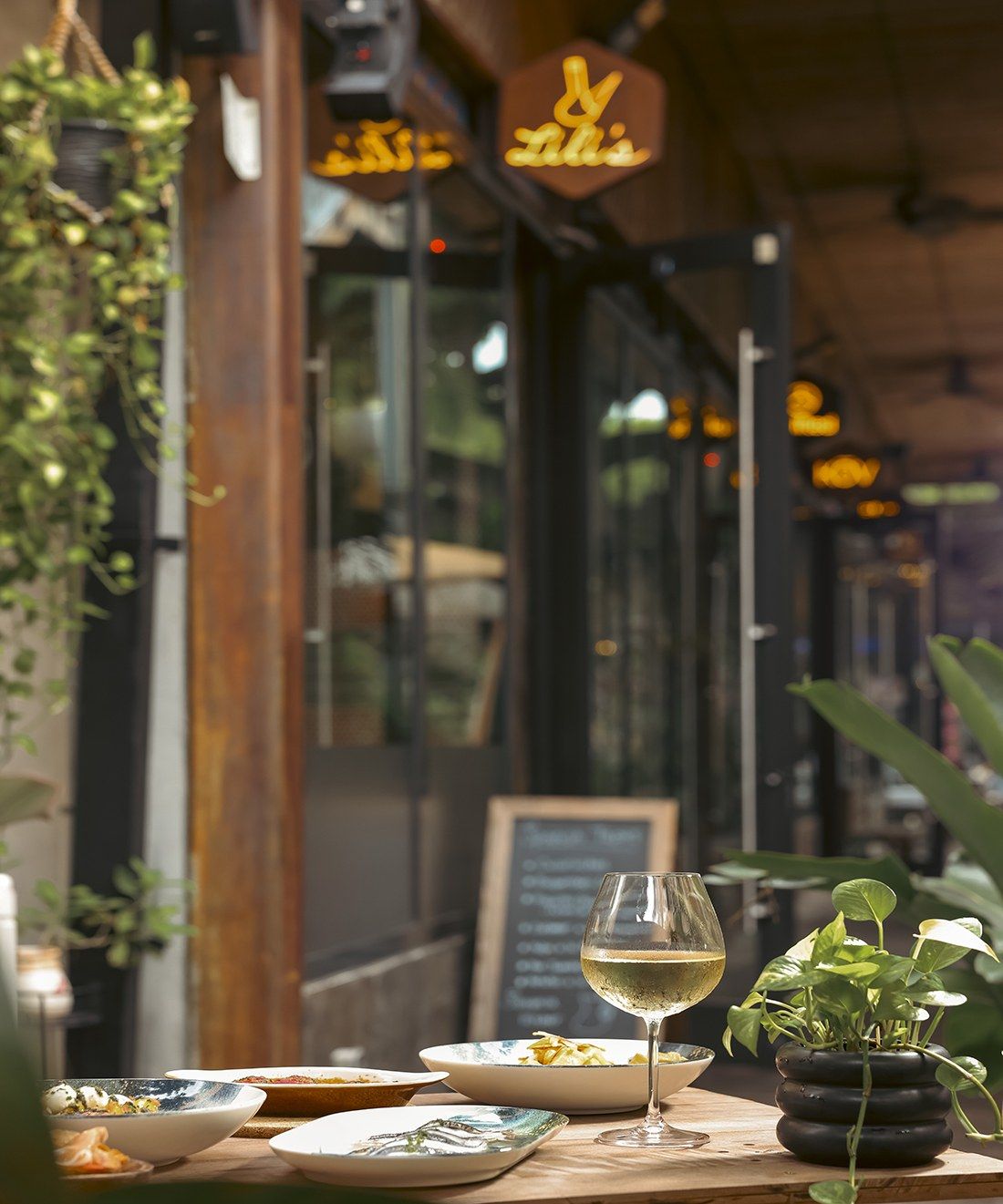 Restaurant storefront with hanging plants and a glass of white wine on a table at dusk