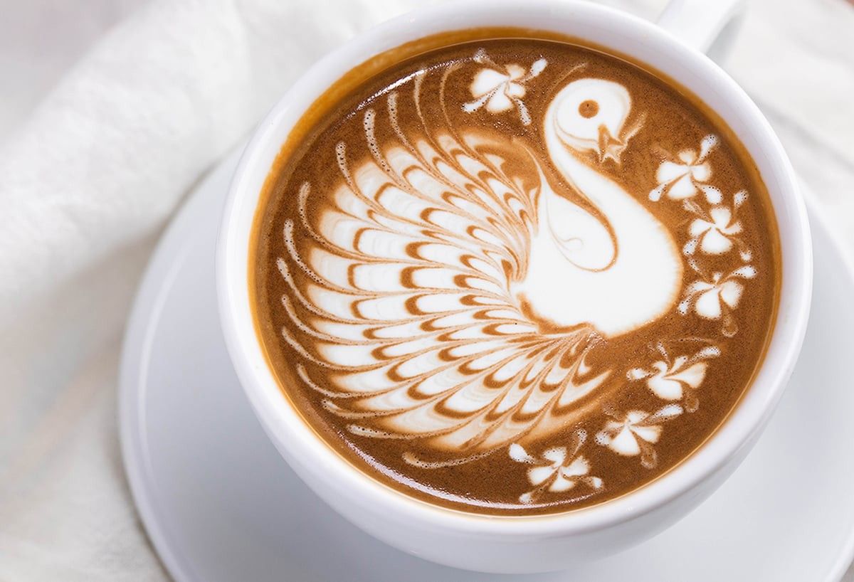 A cup of coffee topped with latte art in the shape of a swan, on a white saucer.