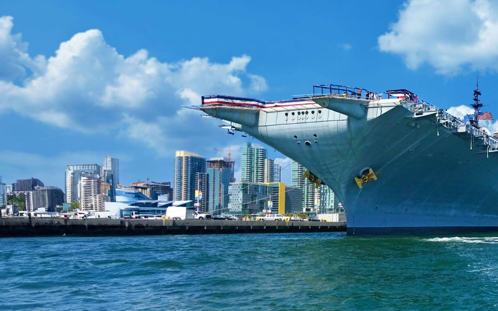 The aircraft carrier docked beside the city skyline under a blue sky with clouds.