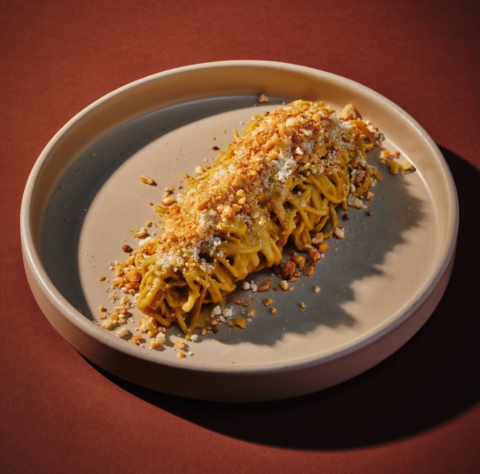 Pasta dish topped with crumbs on a beige plate against a rust-red background