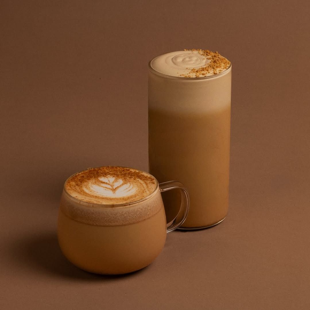 Two lattes, one in a clear glass mug and one in a tall glass, with foam dusted with cinnamon on a brown background.