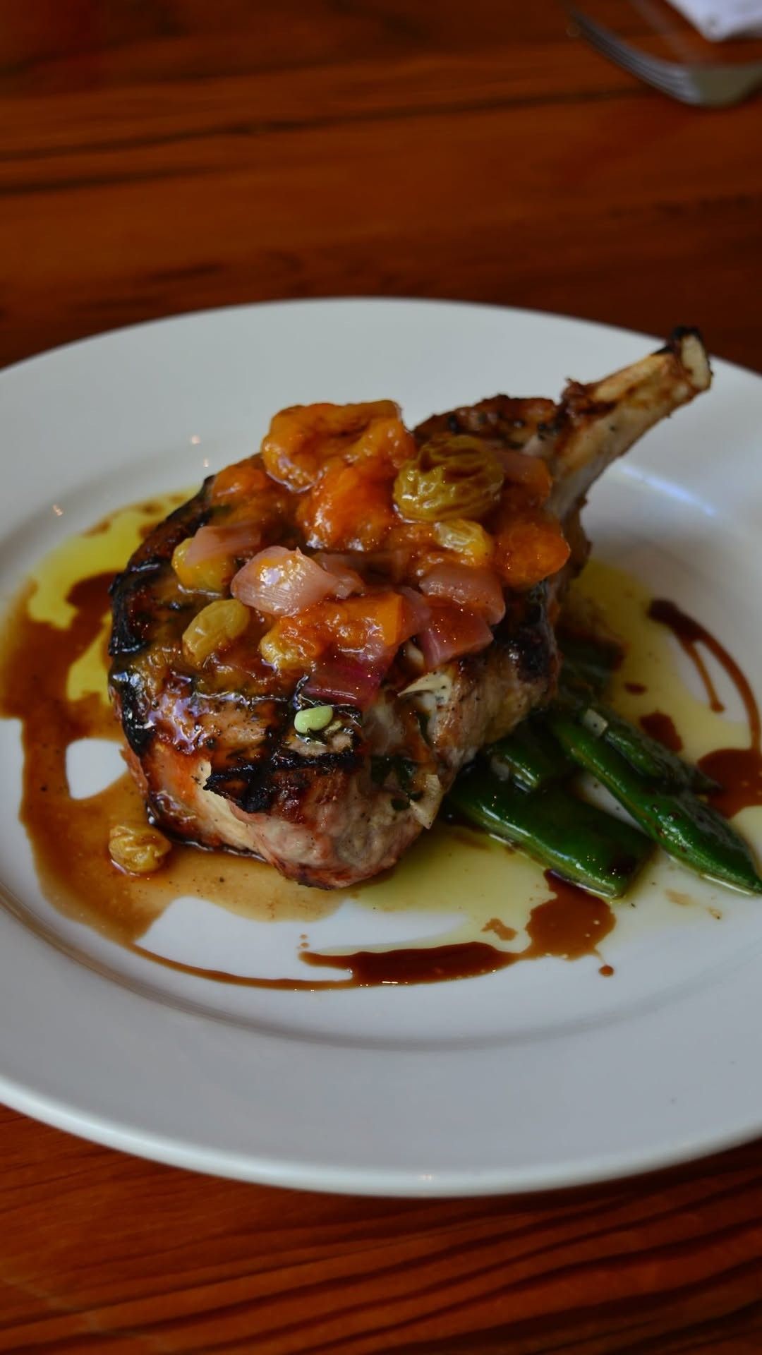 A grilled pork chop topped with a warm tomato and olive relish, with green beans and a drizzle of sauce on a white plate.