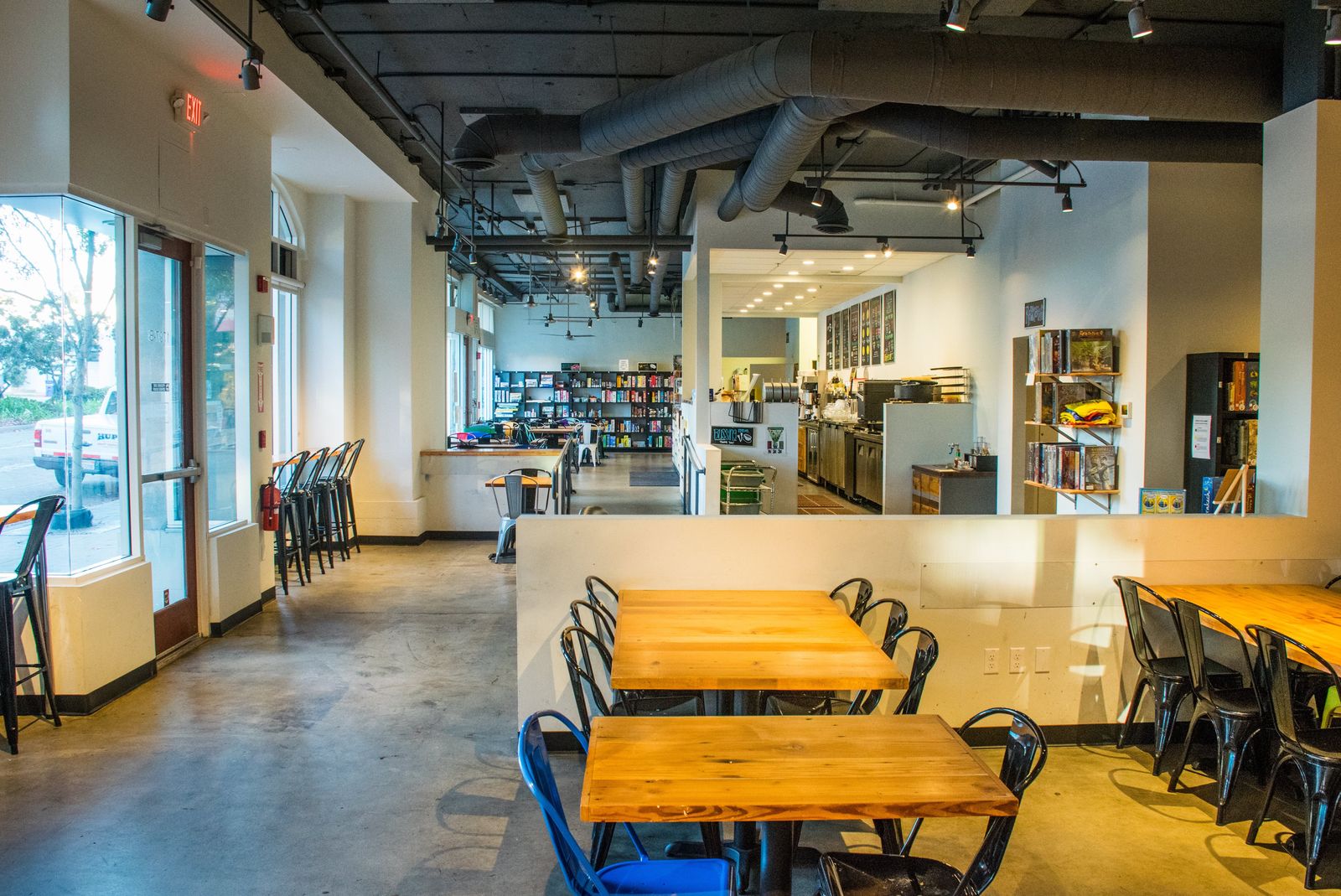 A bright cafe room with wooden tables and black chairs, a counter at the back and large windows along the left wall.