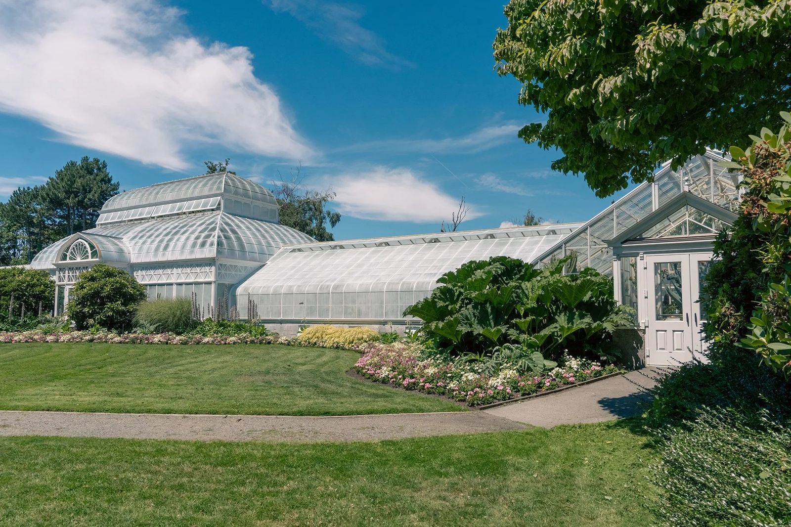 The white glass-and-steel conservatory building behind a lawn and flower beds under a blue sky.