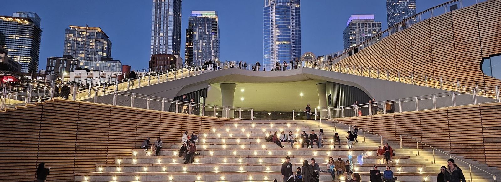 A wide lit staircase leading up to a waterfront promenade with city towers behind it at dusk.