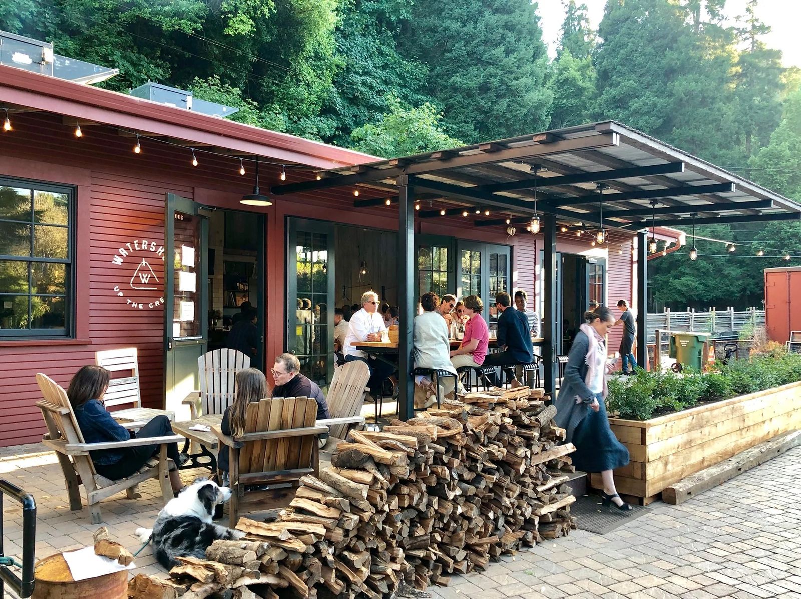 Red wooden building with a covered patio, outdoor seating and stacked firewood among redwoods