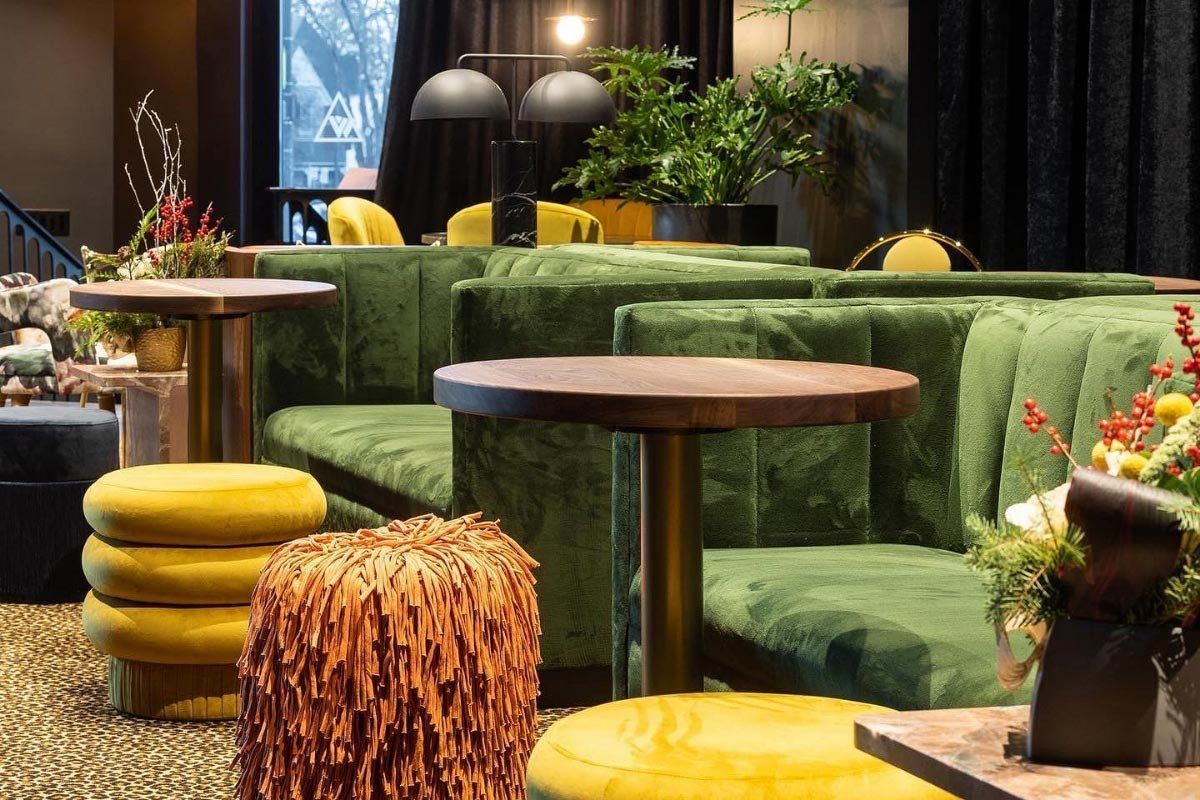 A lounge with green velvet banquettes, round tables, yellow poufs and plants in front of tall windows.