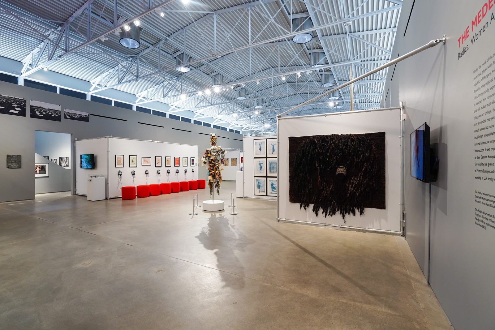 Spacious museum gallery with a steel truss ceiling, framed works on white walls and a red bench