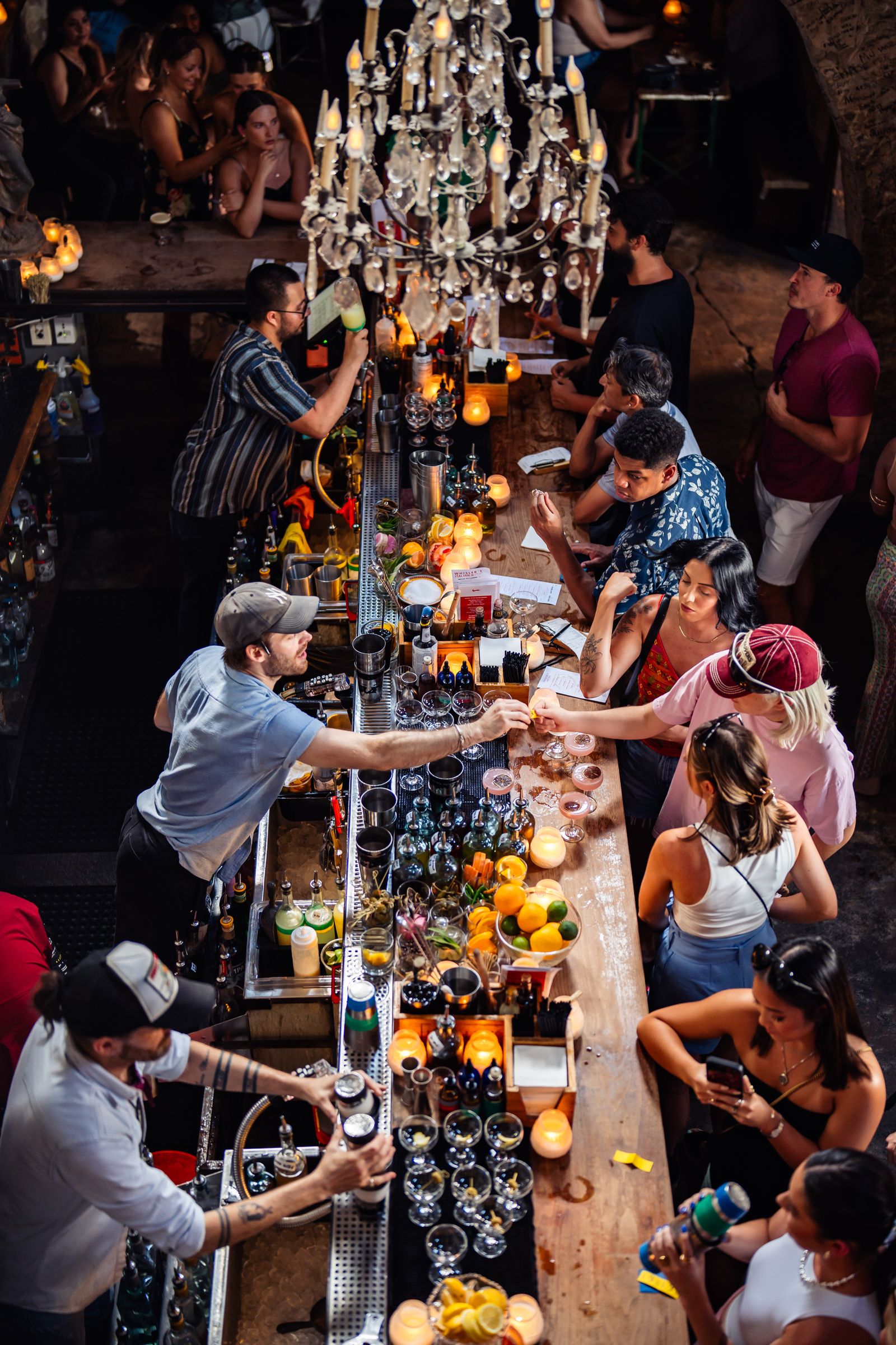 A long bar lit by a chandelier and candles with bartenders and guests along it in a dim room.