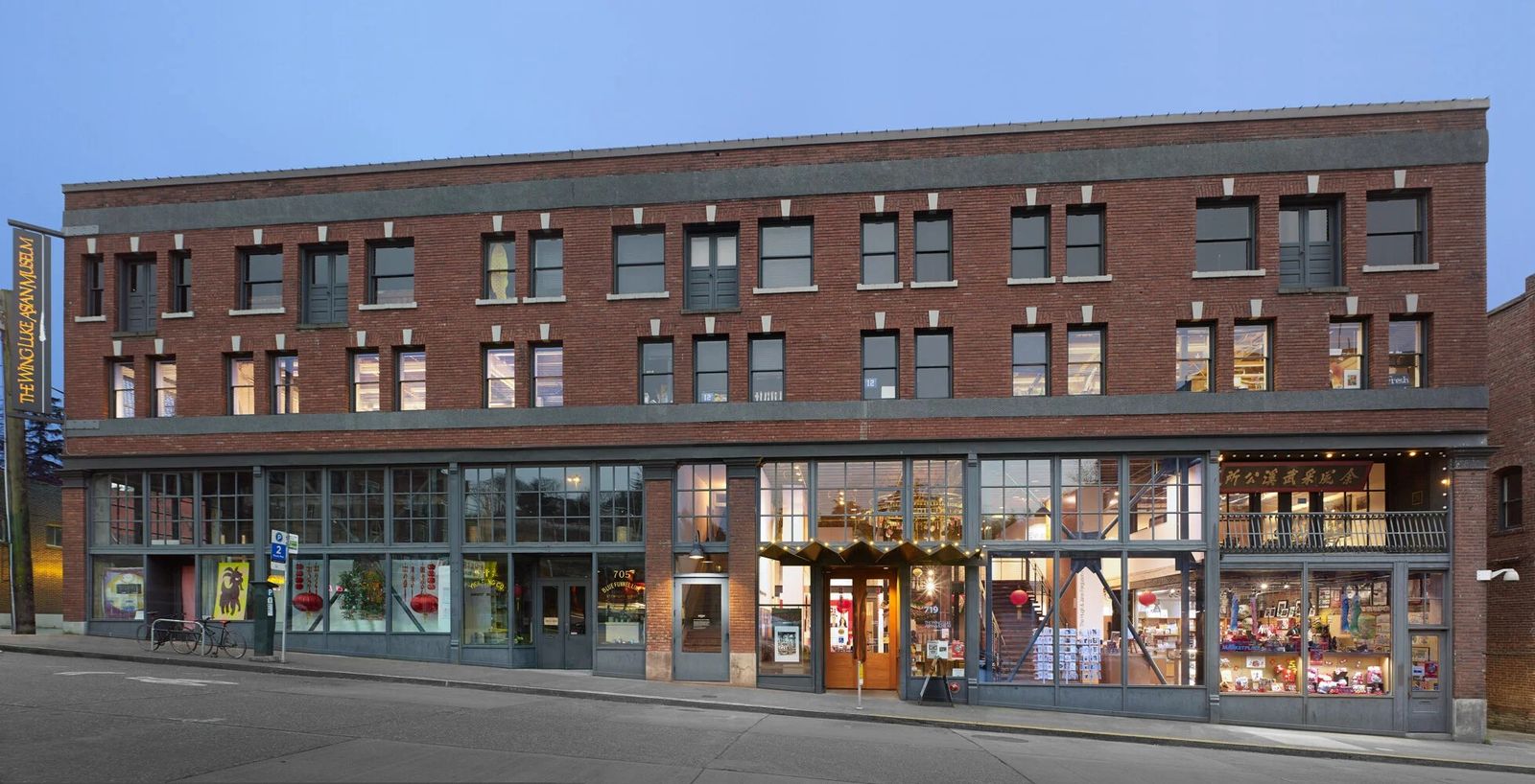 A three-story red brick building with shopfront windows, lit at dusk.