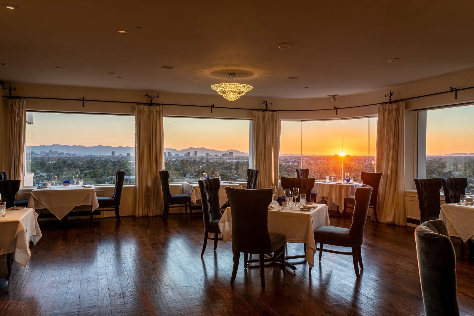 A dining room with set tables and tall windows looking out over the city at sunset