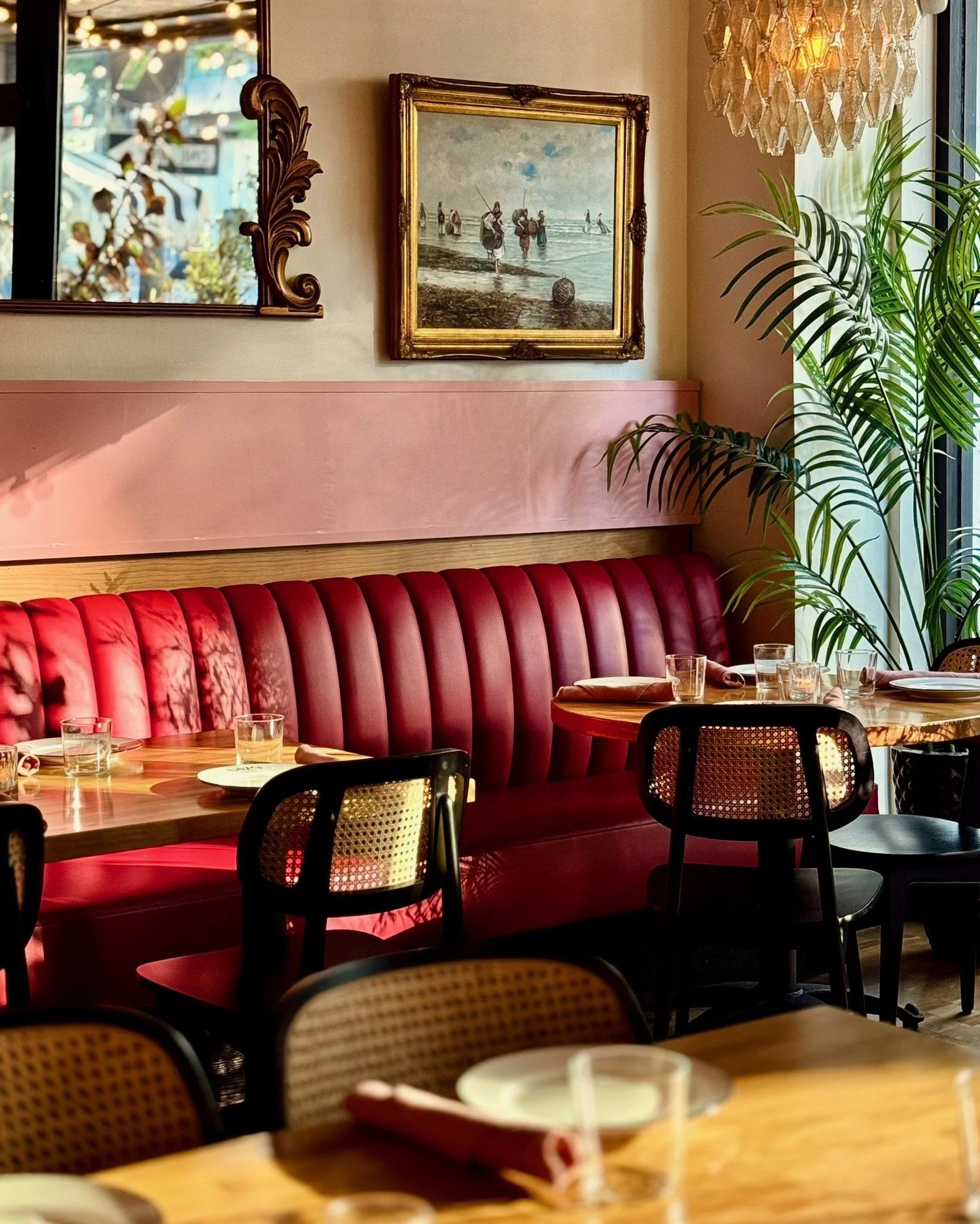 Dining room with a red leather banquette, cane-back chairs, a gilt-framed painting and a palm