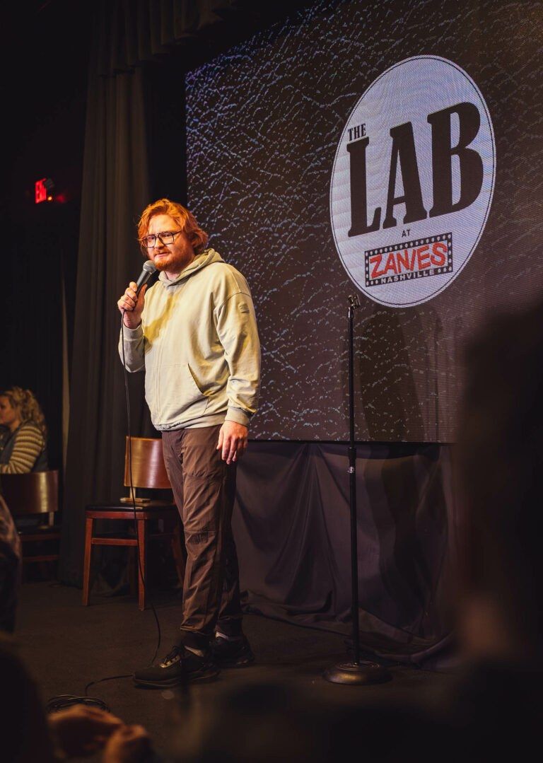 Comedian standing on stage with a microphone in front of The Lab sign
