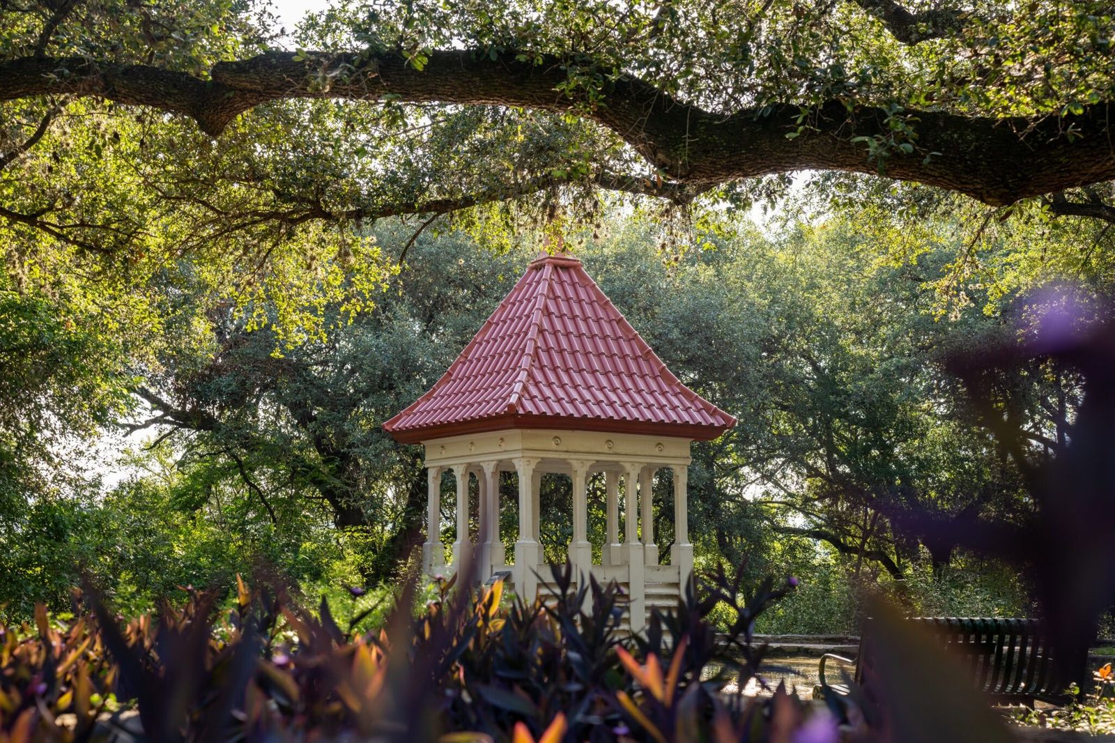 A small garden gazebo with a red roof among live oaks and purple flowers.