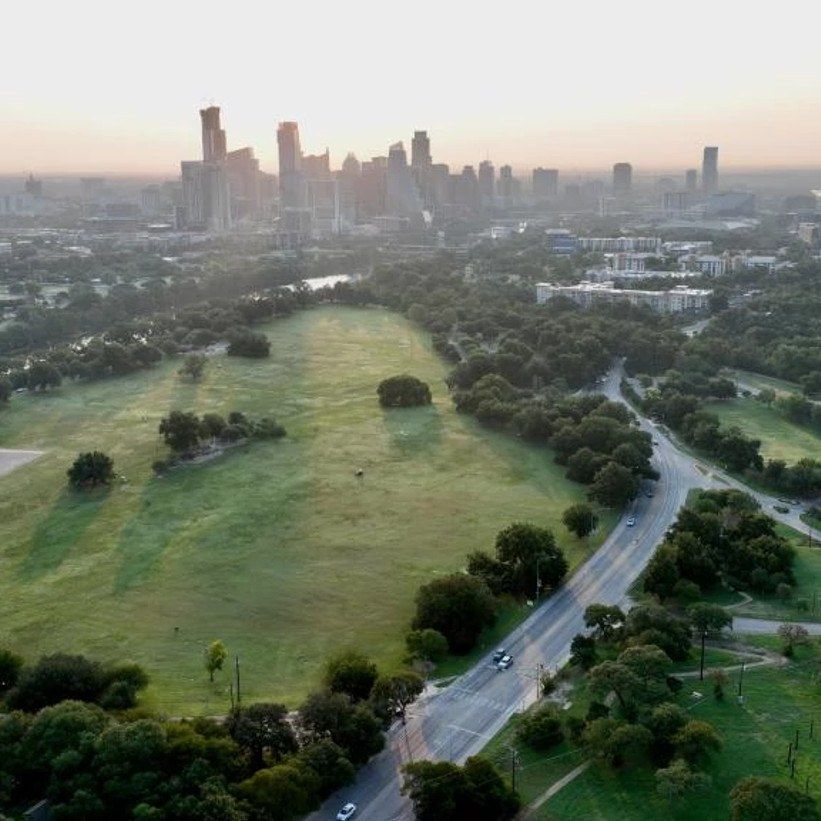 An aerial view of a green lawn and winding road under trees with the Austin skyline in the distance.