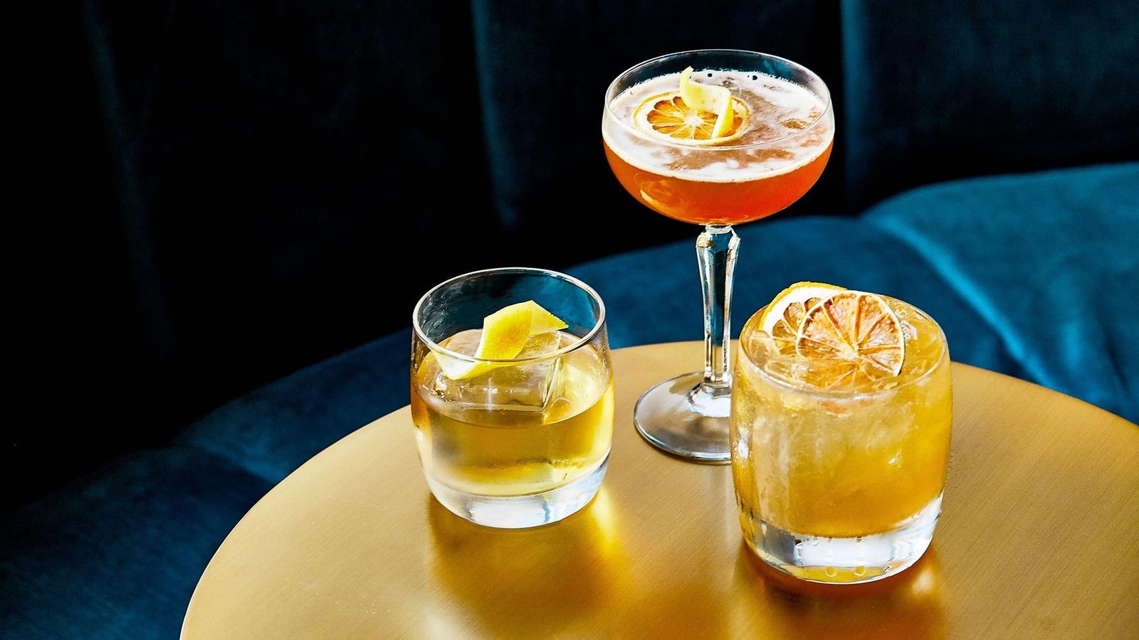 Two cocktails, one in a coupe garnished with citrus, on a round gold table.